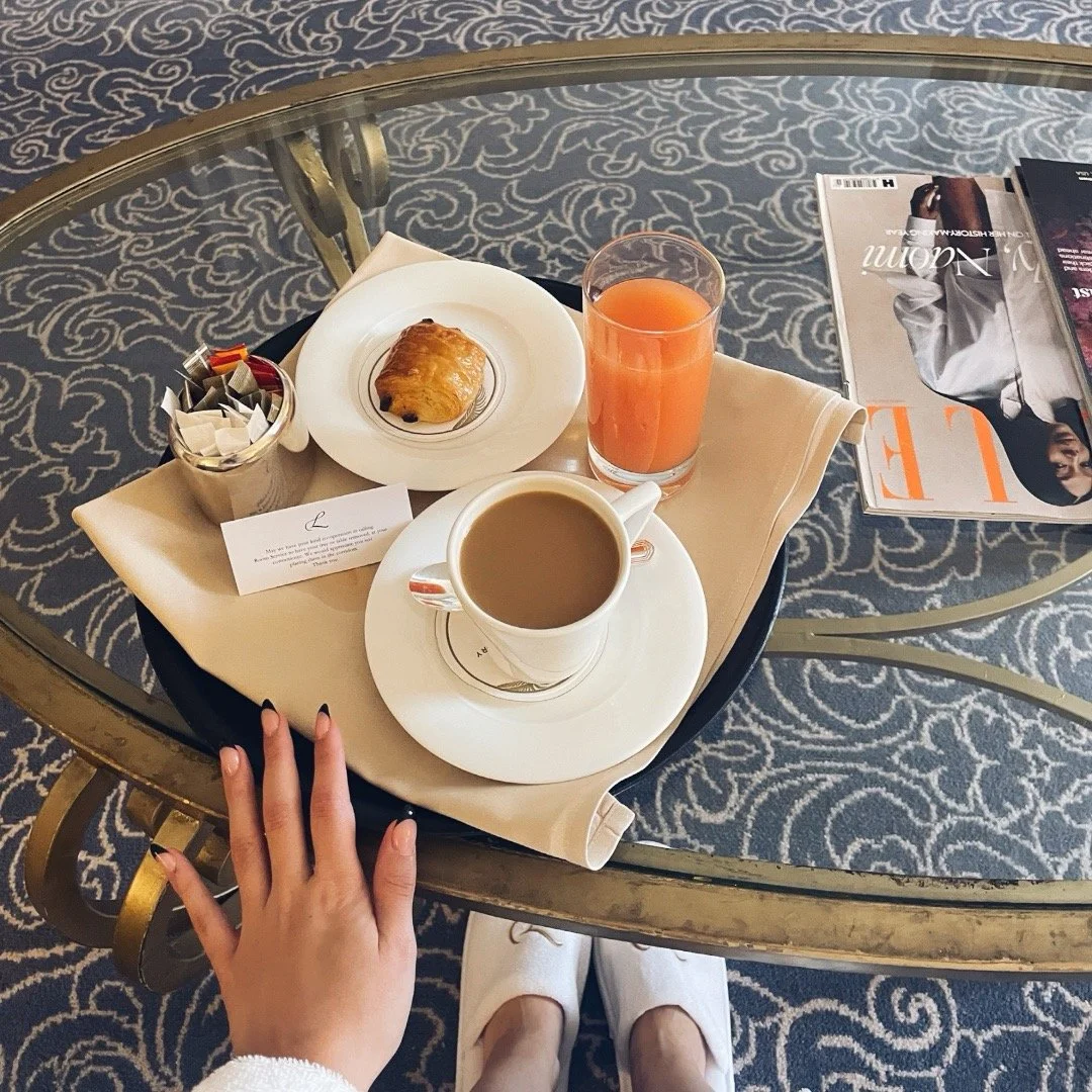 Breakfast tray with coffee, orange juice, a croissant, and a sugar packet, with a magazine and a hand with painted nails visible in a hotel or lounge setting.