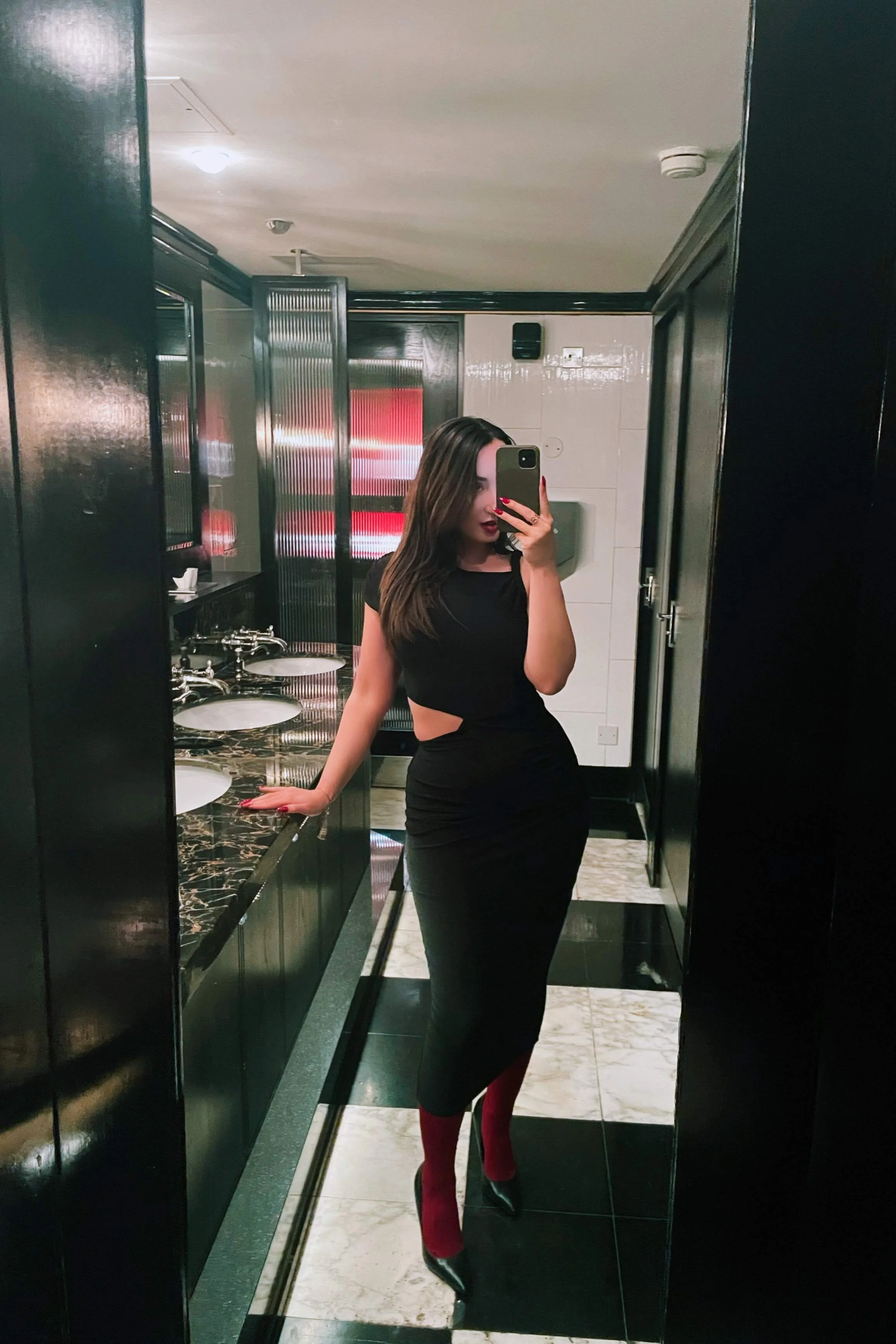 A woman in a black dress with cutouts on the side, red socks, and black high heels taking a mirror selfie in a public restroom with a black and white tiled floor and sinks with black marble countertops.