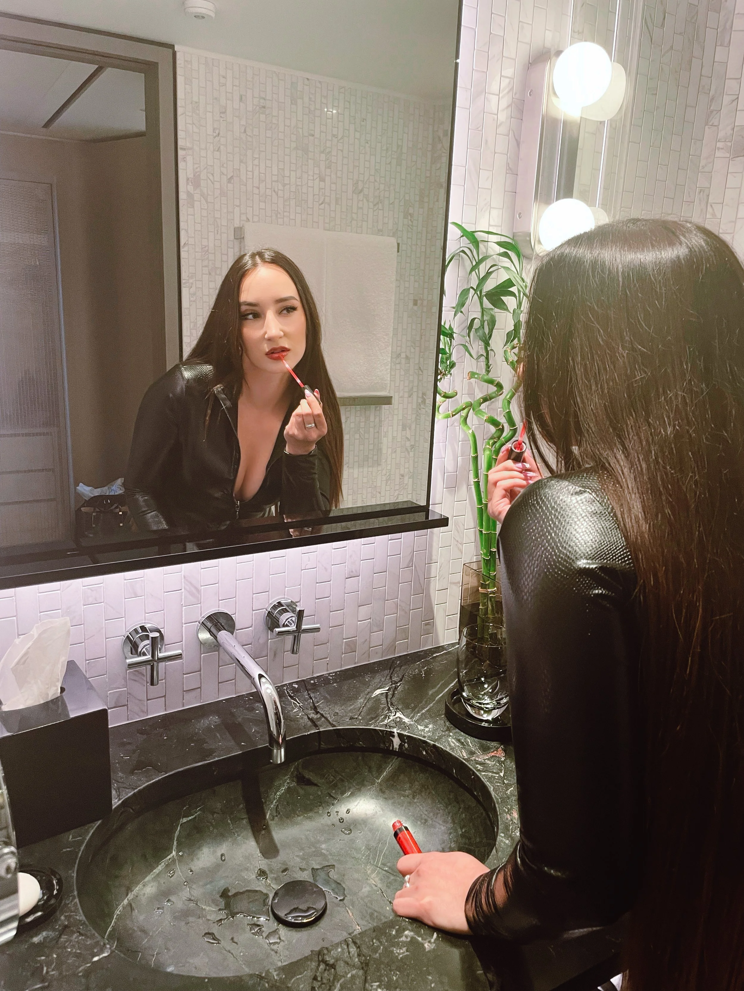 A woman in a black jacket is applying lipstick in front of a mirror in a modern bathroom with a black marble sink, a tissue box, a green plant, and round vanity lights.