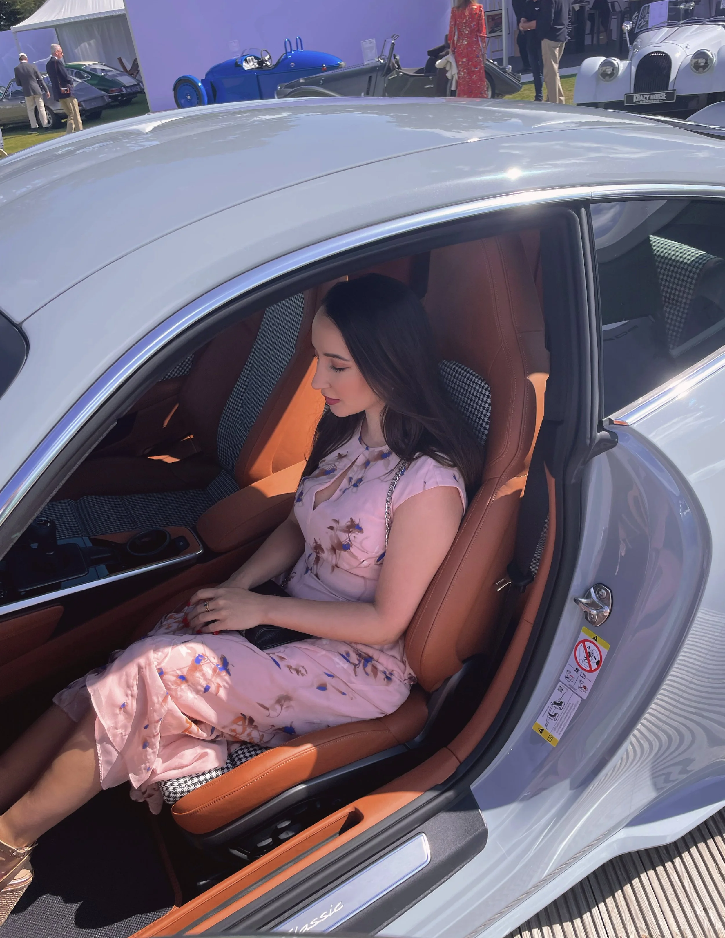 Woman in pink floral dress sitting inside a luxury car with tan leather interior at an auto show.