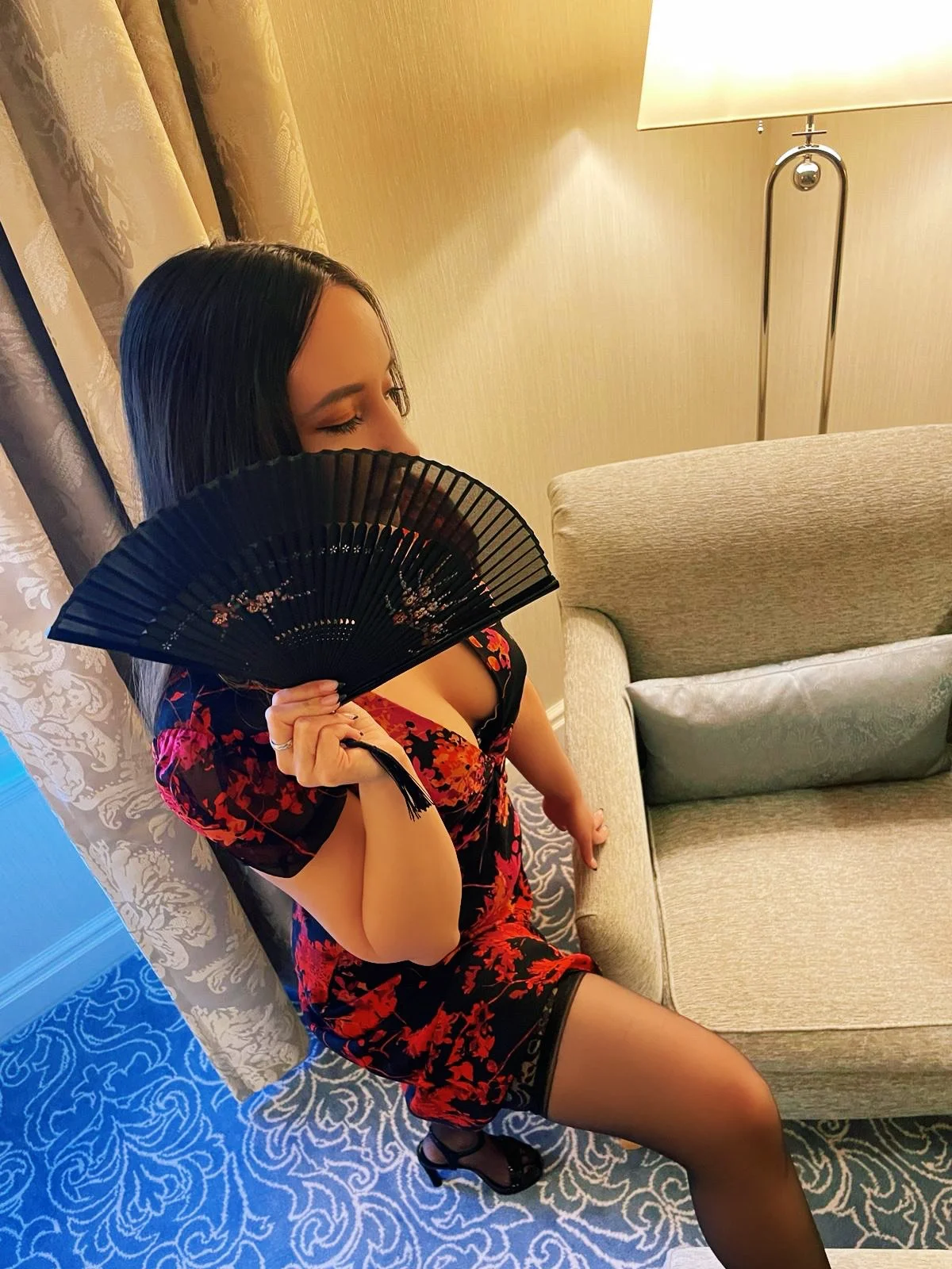 Woman holding a black fan, dressed in a black and red floral-patterned dress, standing next to an armchair in a hotel room.