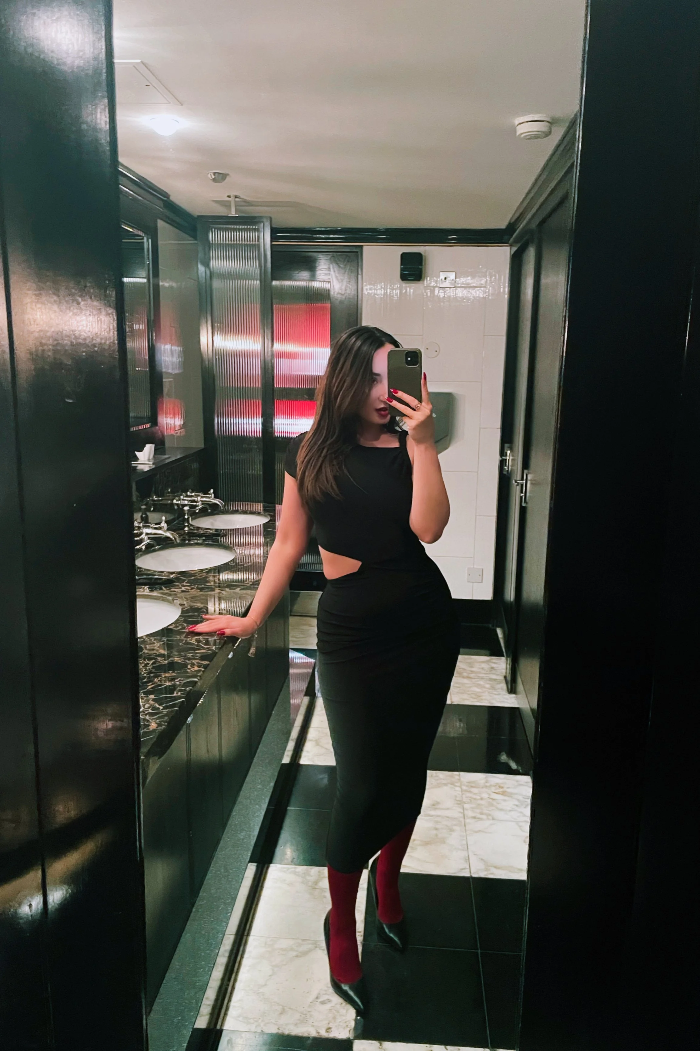 A woman taking a selfie in a restroom mirror, wearing a black dress with side cutouts, red tights, and black heels.