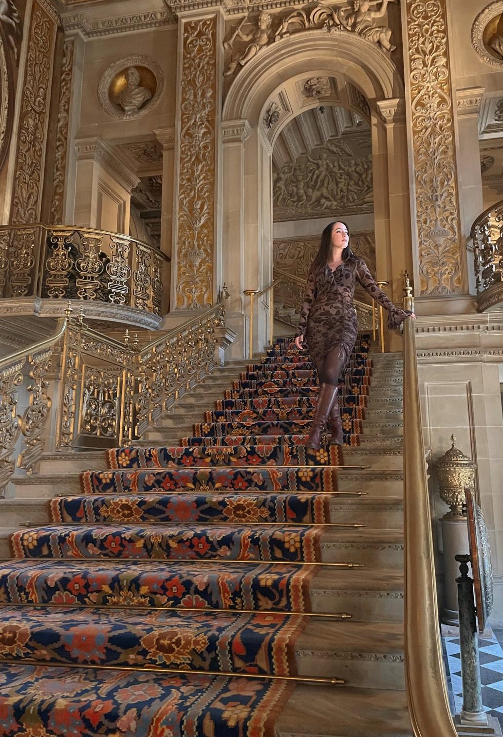 Woman standing on a grand staircase in an ornate, gold-decorated interior with detailed carvings, statues, and a patterned carpet.
