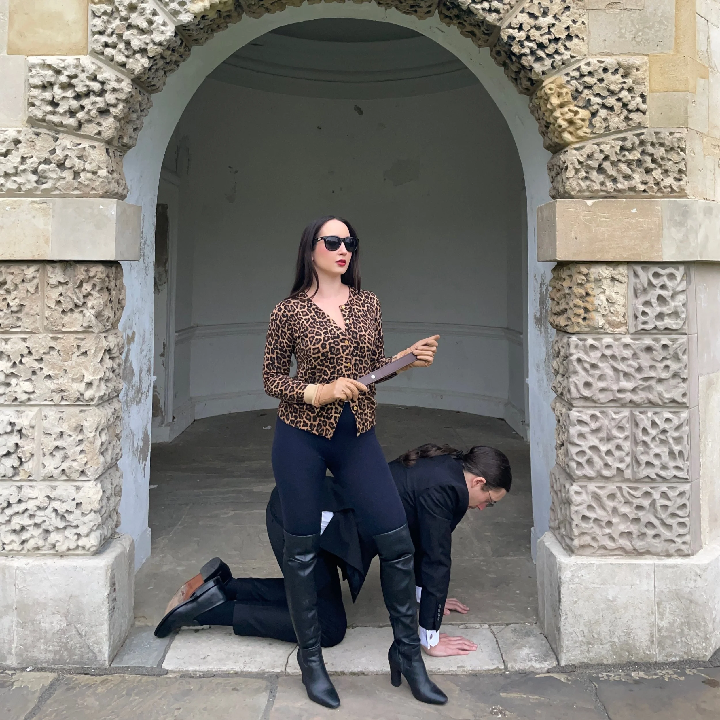 A woman in a leopard print top and black boots stands with one foot on a person kneeling on the ground beneath her. The person kneeling is dressed in a black suit and appears to be in a praying position with hands on the ground. They are under an arc