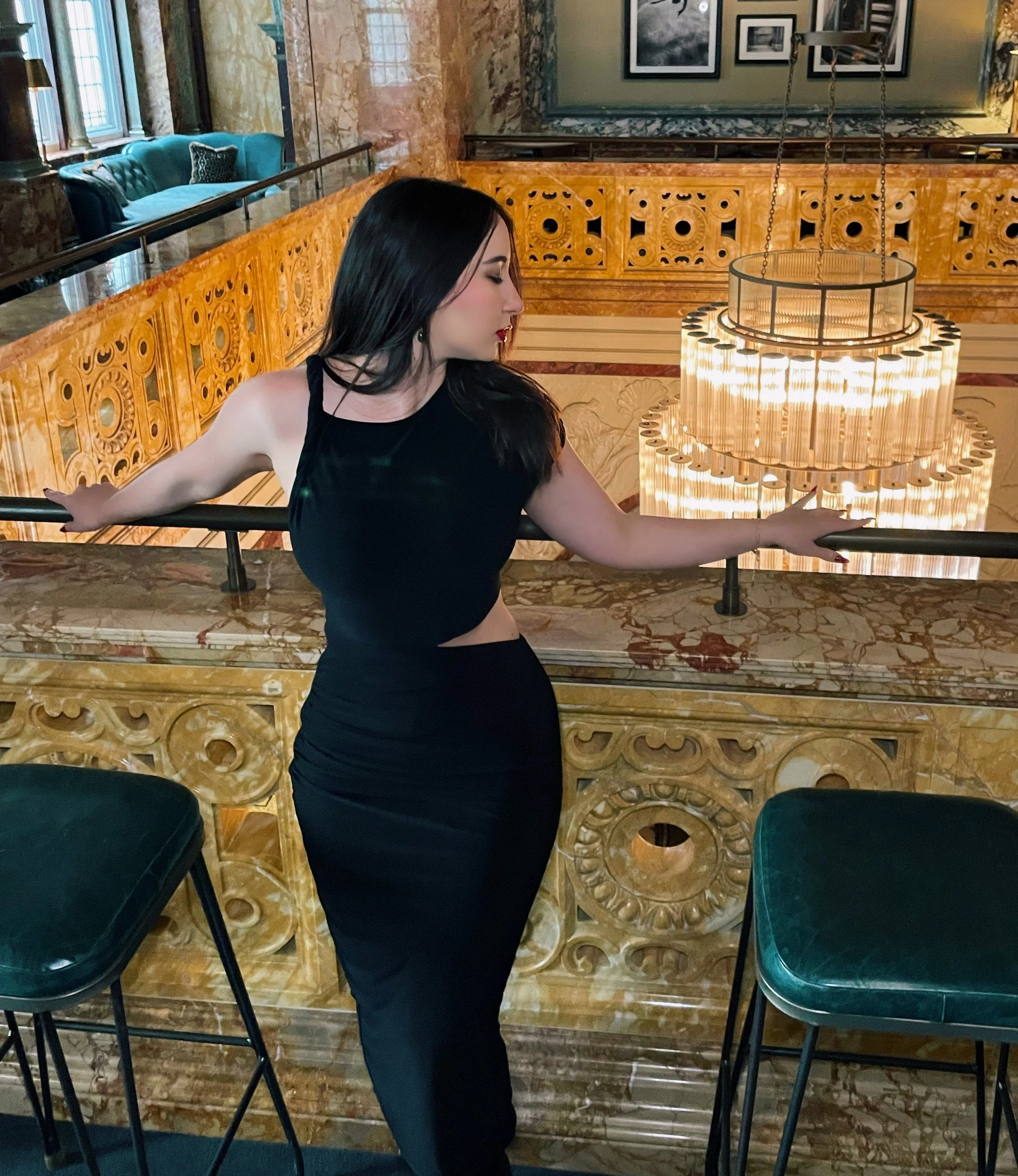 High-class Escort with dark hair in a black dress standing with arms outstretched, holding onto a railing, in an elegant indoor setting with ornate yellow and marble decor, staircases, and a large chandelier in the background.