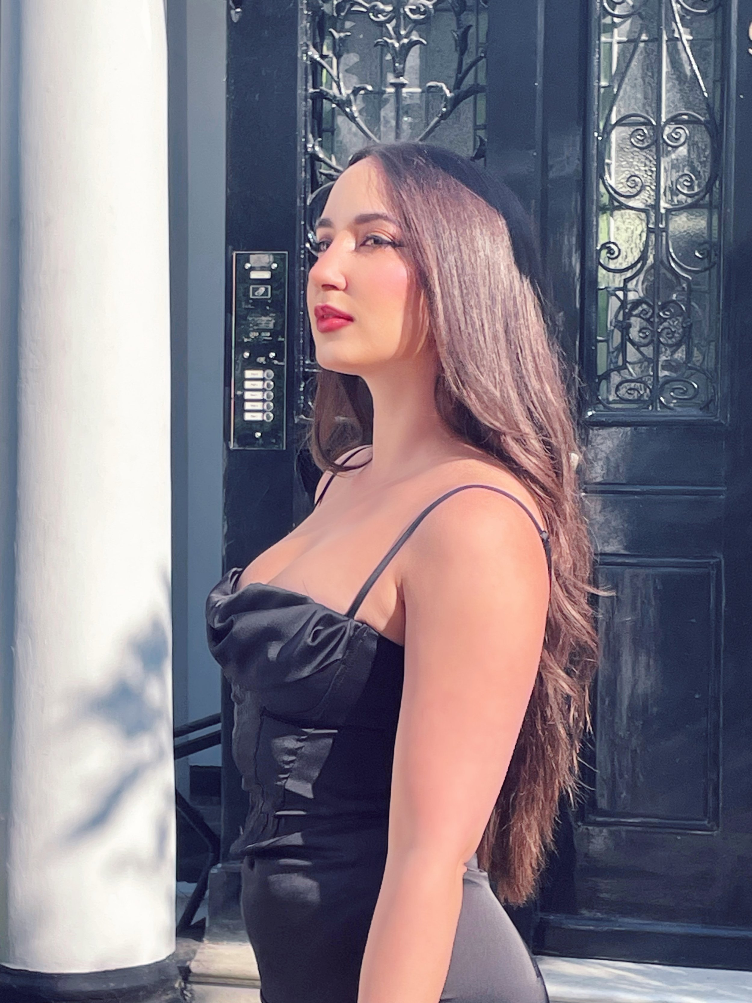 A woman with long, wavy brown hair standing in front of a wrought iron gate and black door, wearing a black spaghetti strap top.