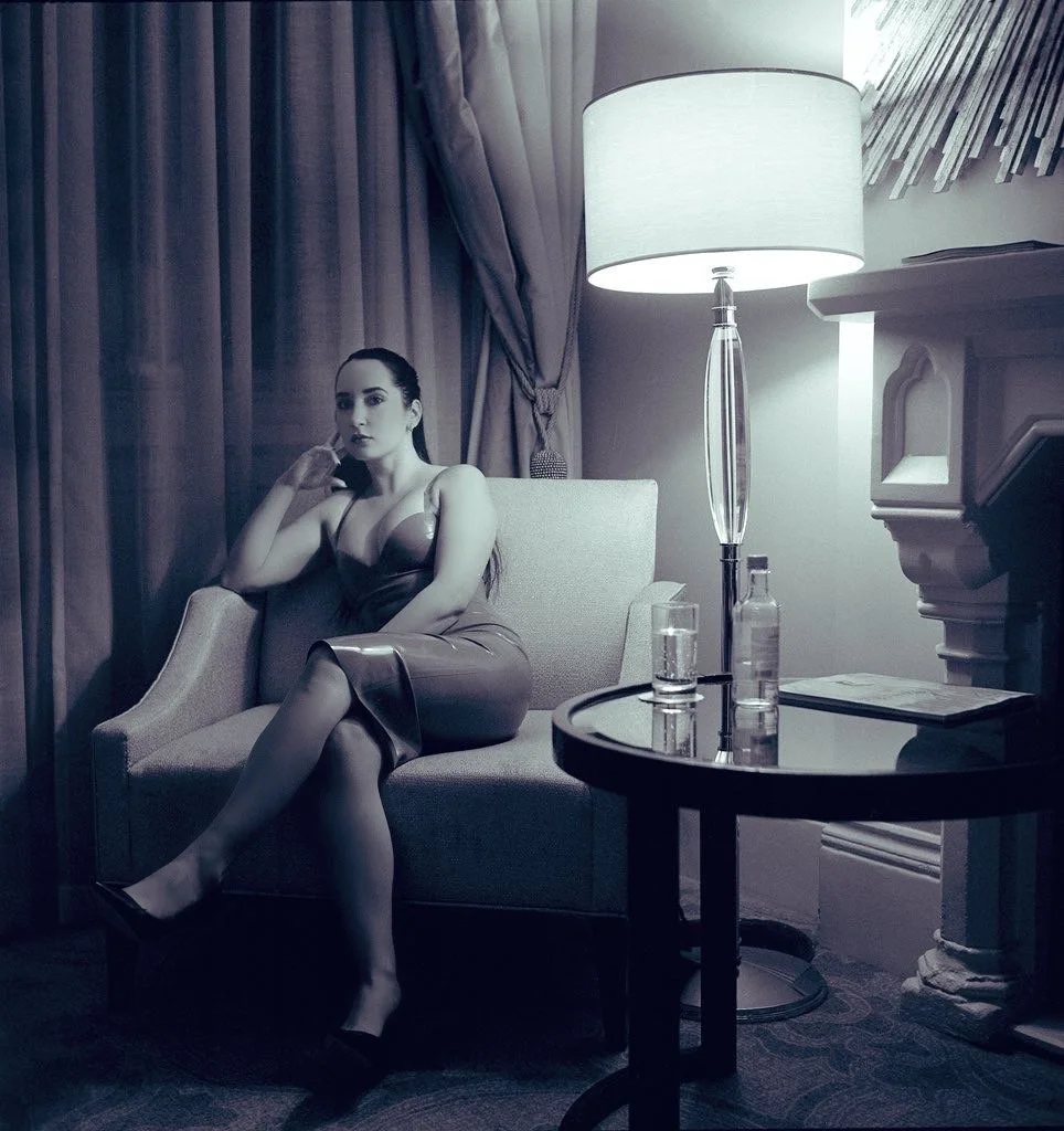 A woman in a satin dress sitting on a sofa in a dimly lit room with a lamp on a table nearby.