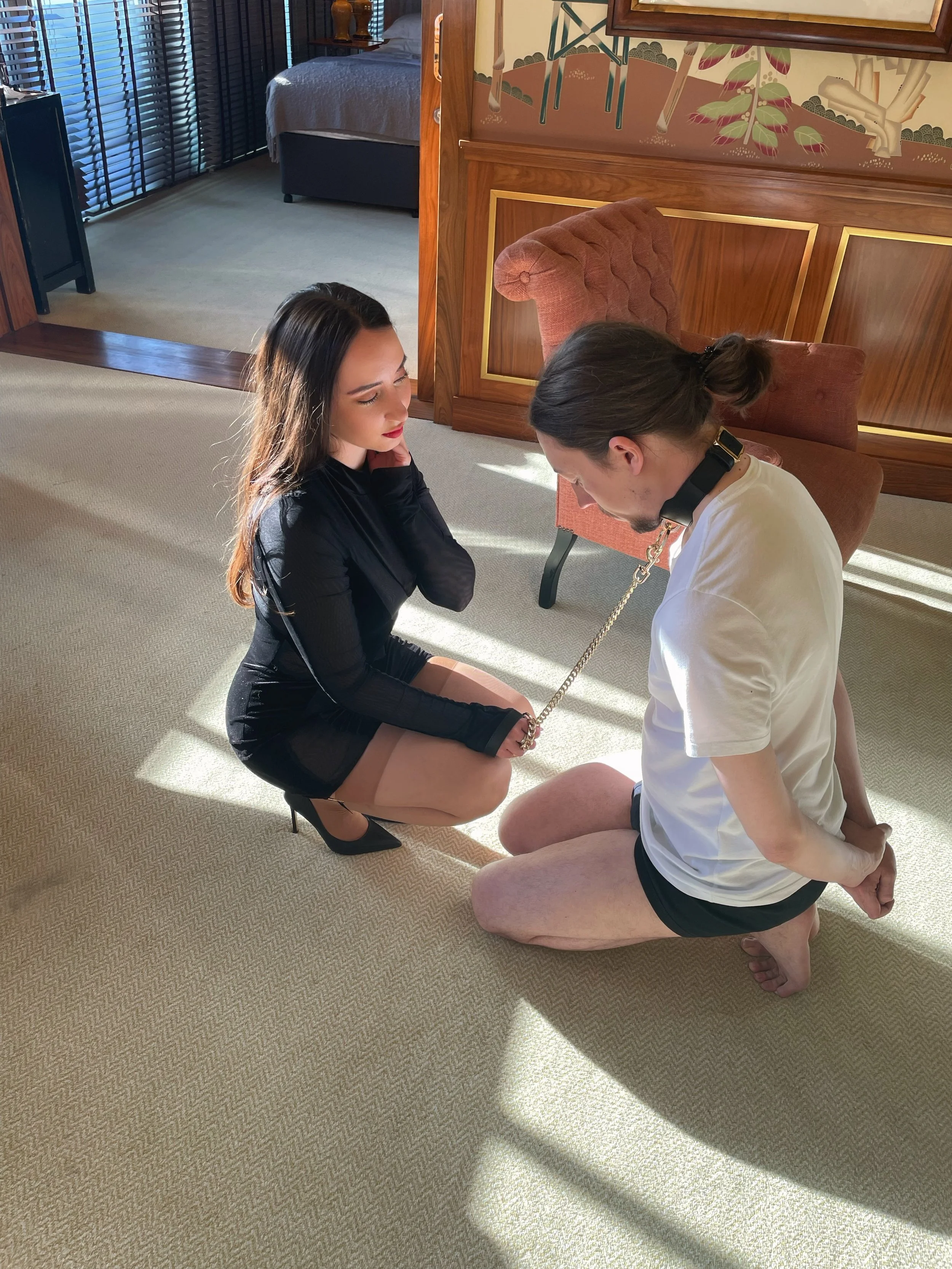 A woman in black high heels and a sheer black dress kneels on a beige carpet, holding a chain leash attached to a man kneeling and dressed in a white t-shirt and black shorts. They are indoors with sunlight and shadows on the carpet, and a vintage-st