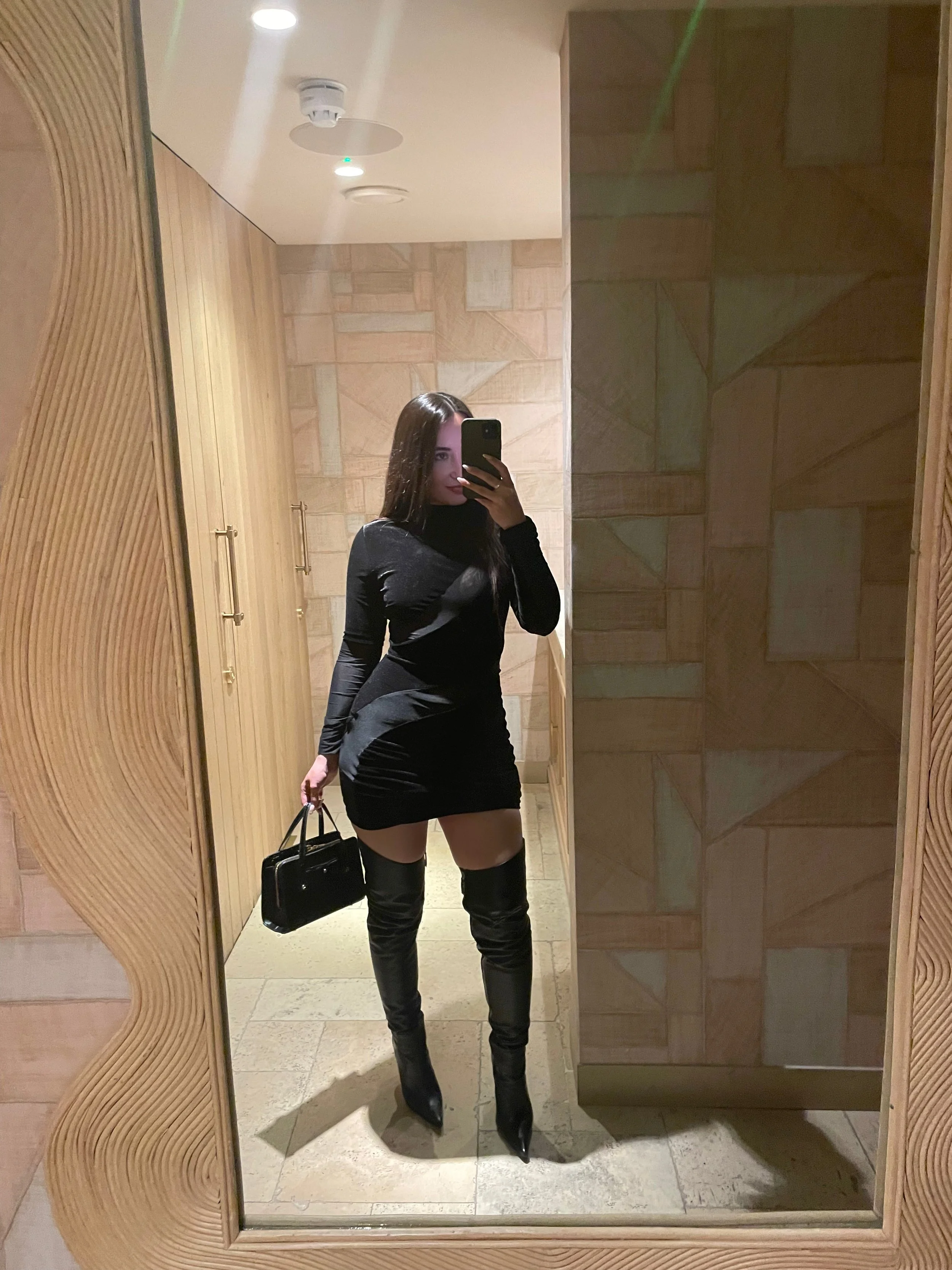 A woman in a black dress and over-the-knee black boots taking a mirror selfie in a room with wooden wall decor, holding a black handbag.