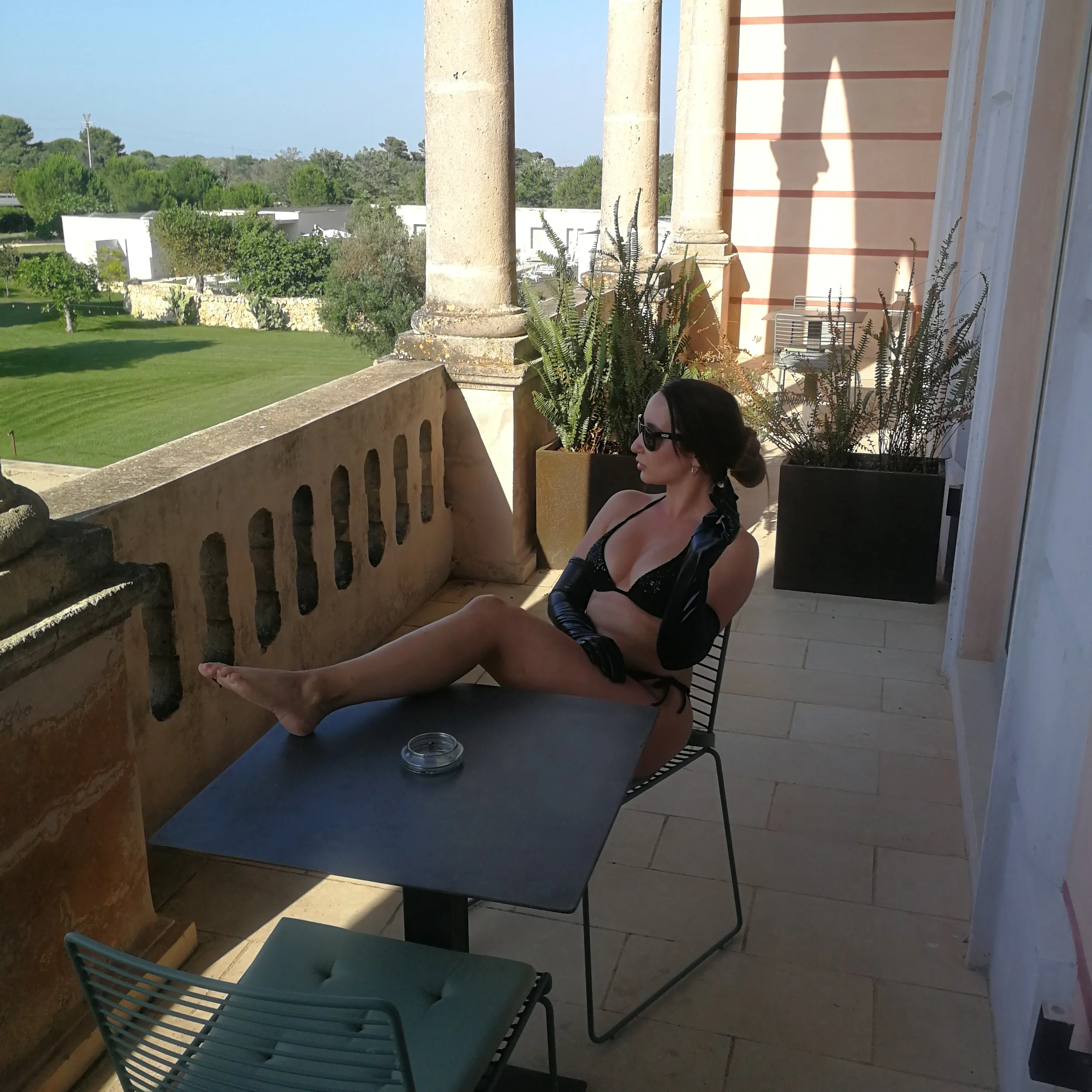 A woman with dark hair wearing sunglasses, a black lingerie top, and long black gloves, sitting on a chair on a balcony with a scenic view of greenery and modern buildings in the background.
