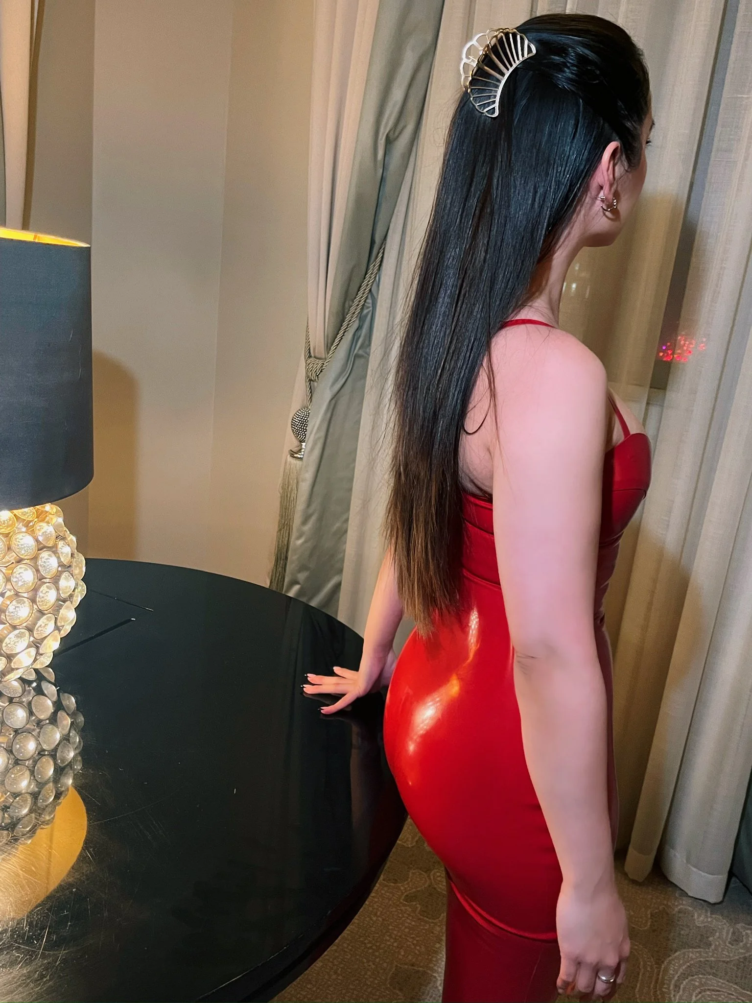 A woman with long black hair dressed in a tight, shiny red dress stands next to a black table, with her hand resting on it, in a room with beige curtains and a lamp with a decorative base.