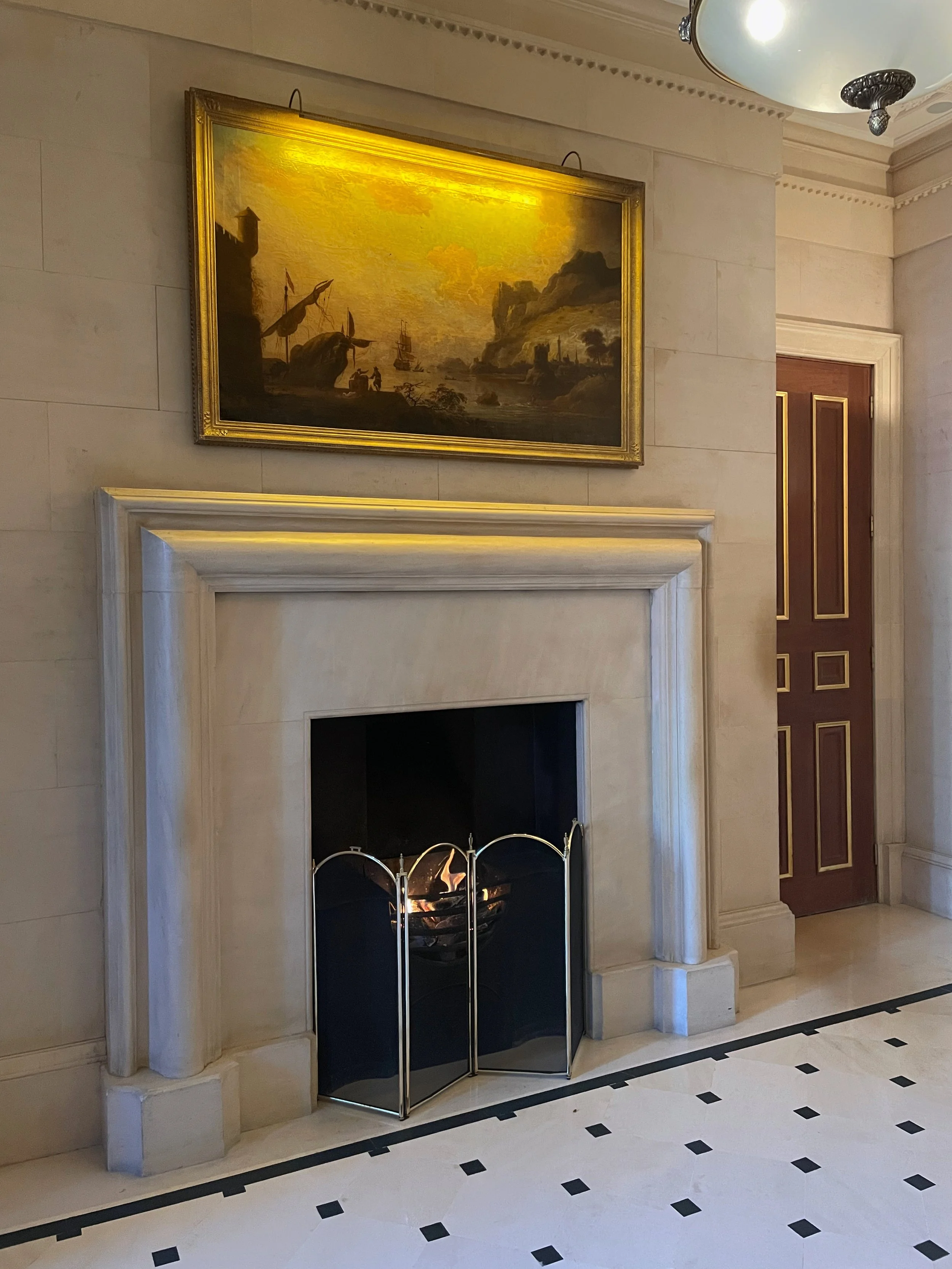 Interior of a room with a lit fireplace, a painting of a harbor scene hanging above the mantel, a wooden door with gold trim to the right, and a ceiling light fixture.