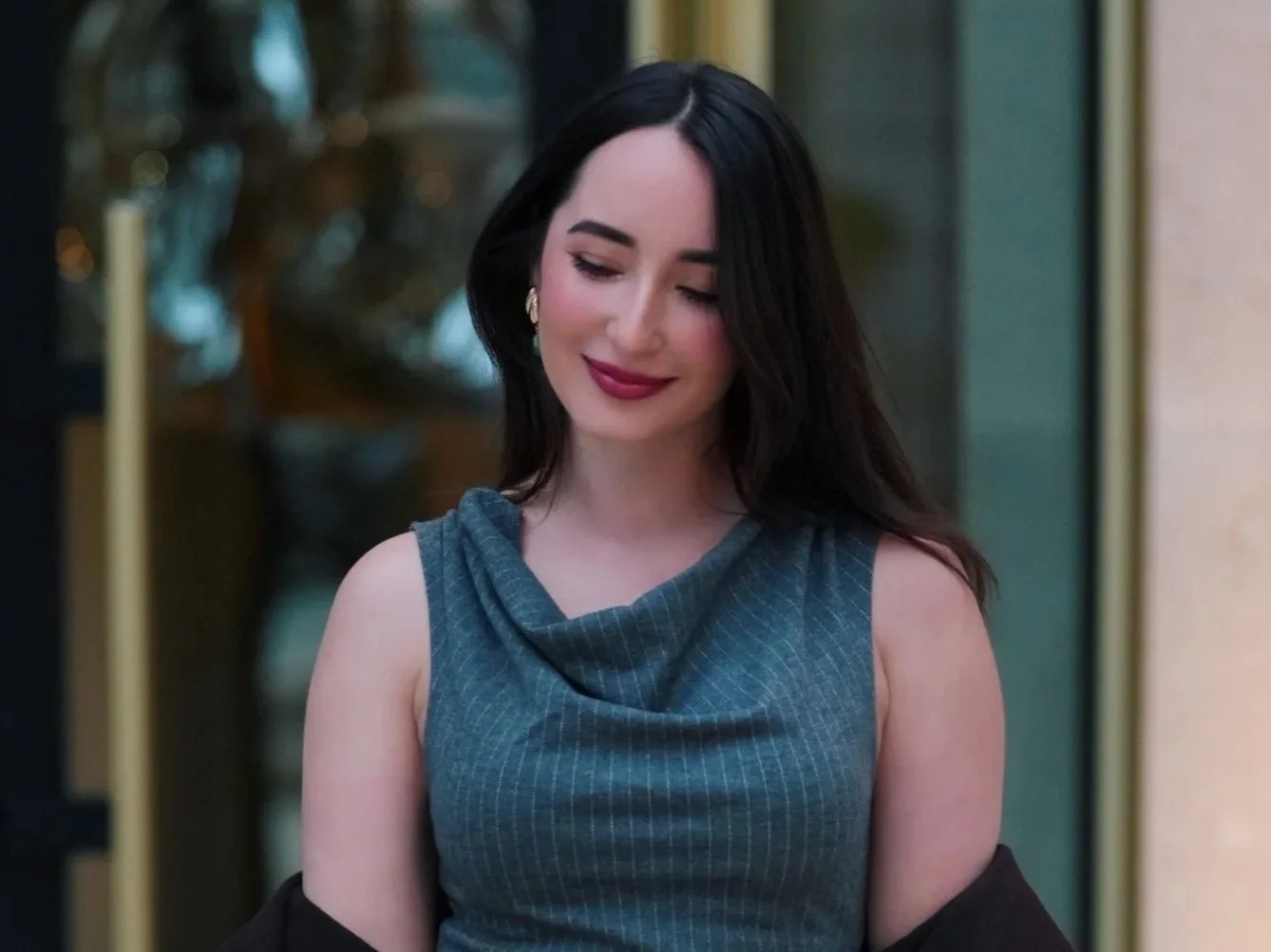 A woman with long dark hair and fair skin is smiling with her eyes closed. She is wearing a sleeveless, blue, cowl-neck top and gold earrings. Behind her is a blurred background.