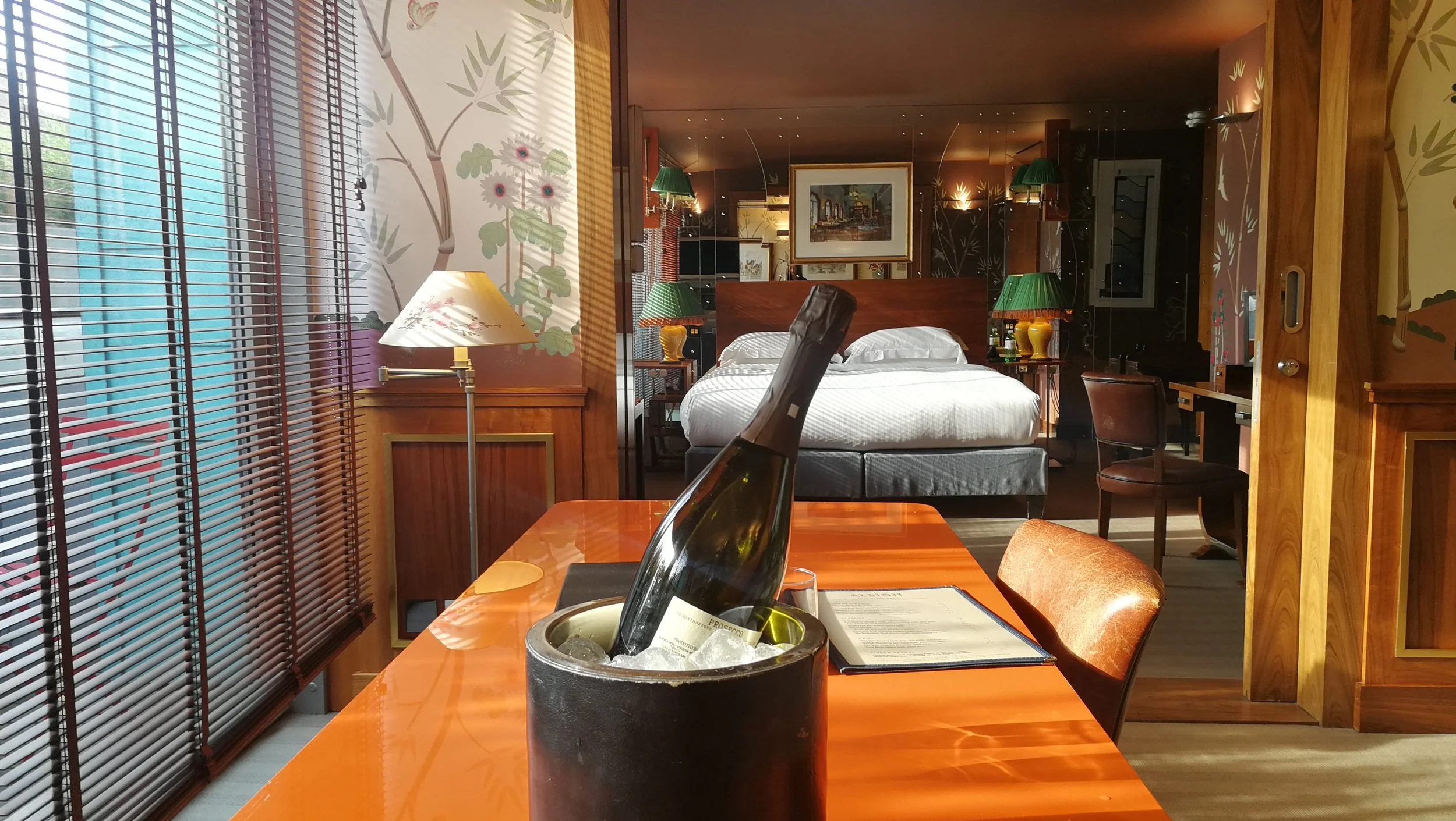 Hotel room with a bed, nightstands, lamps, and artwork, viewed from a table with a wine bottle in an ice bucket and a menu in the foreground.