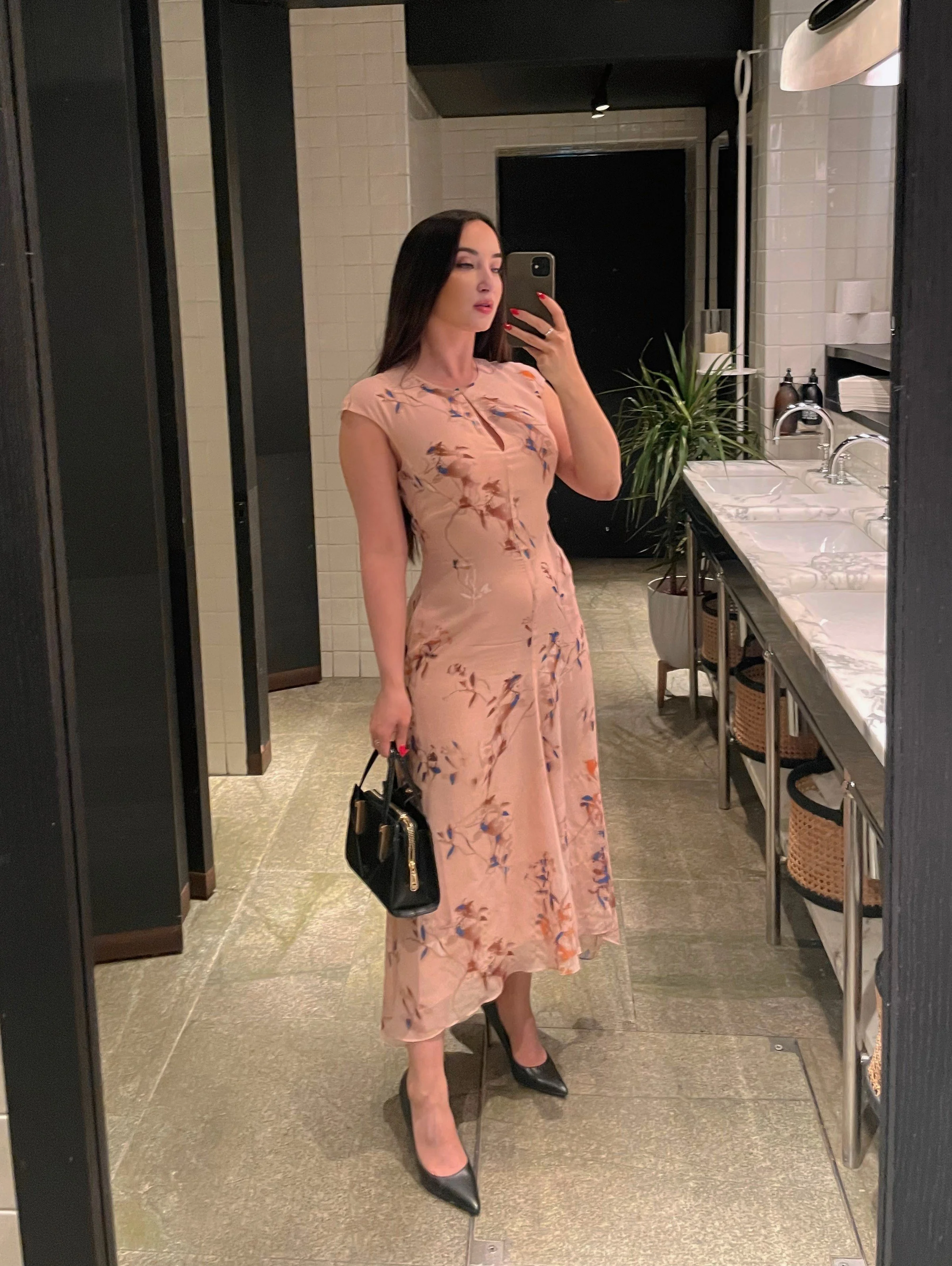 A woman taking a mirror selfie in a restroom. She is wearing a blush pink floral dress with black high heels and holding a black handbag. The restroom has white tiled walls, a countertop with sinks, and a large mirror.