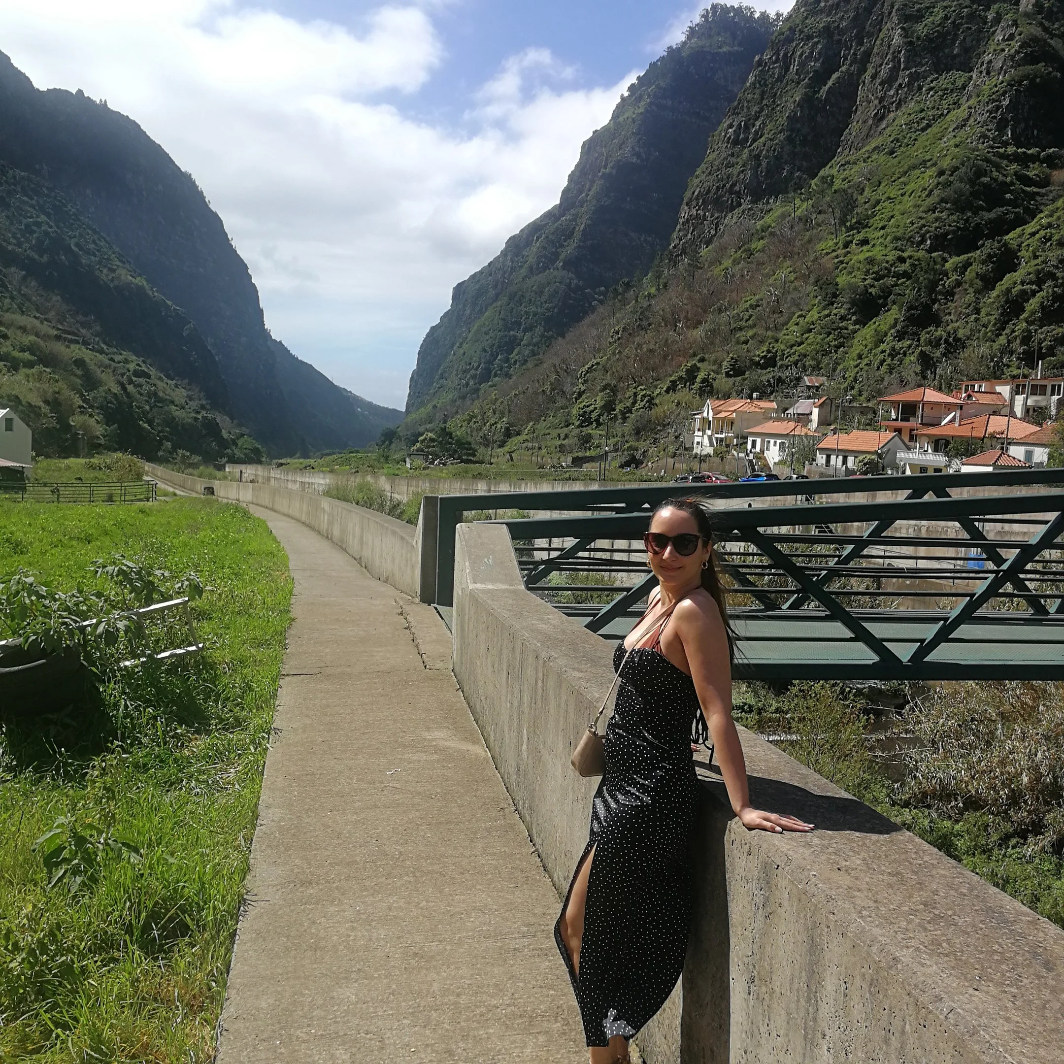 Woman in a black dress with white polka dots wearing sunglasses, standing by a concrete barrier on a bridge with a scenic view of green mountains and a small village.
