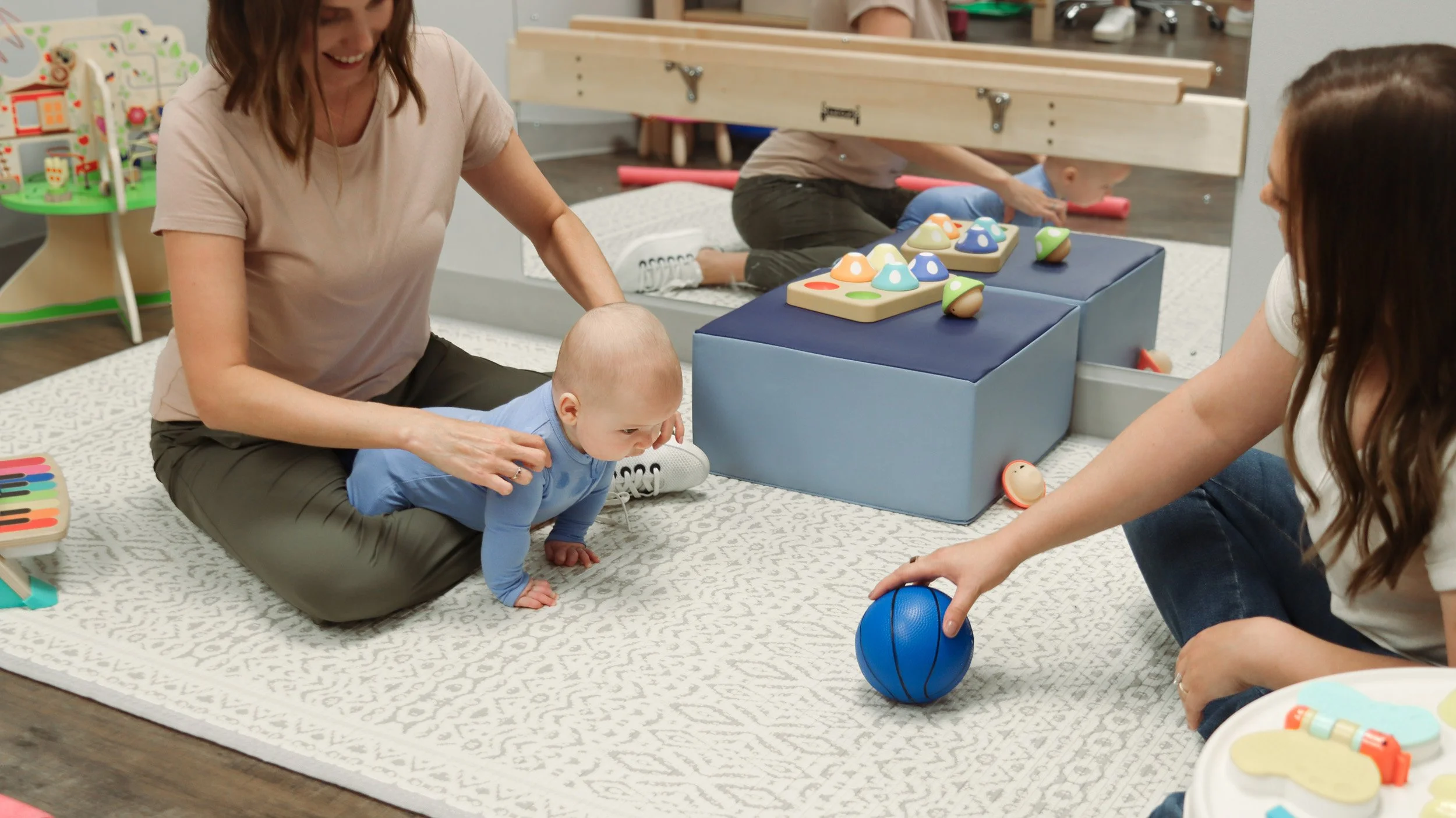 A baby does tummy time exercises with a blue ball, as a pediatric physical therapist positions him correctly