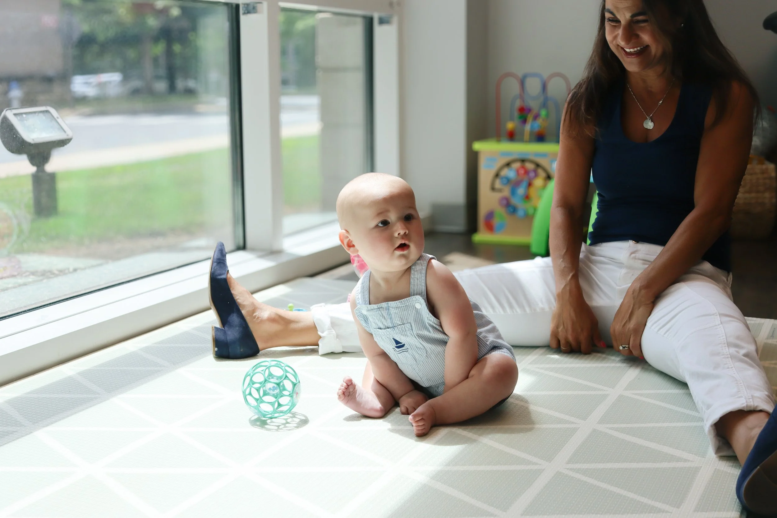 A baby sits and leans toward a toy while Andrea Callanen, owner of Looking Ahead Pediatric Therapies, looks on