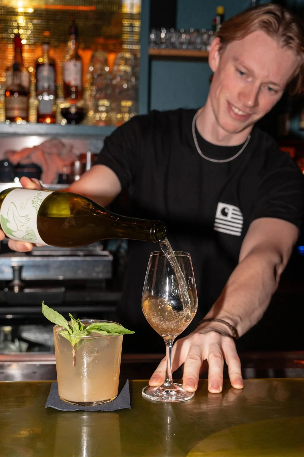 A bartender is pouring a drink into a wine glass, with a cocktail garnished with fresh green herbs on the counter in front.