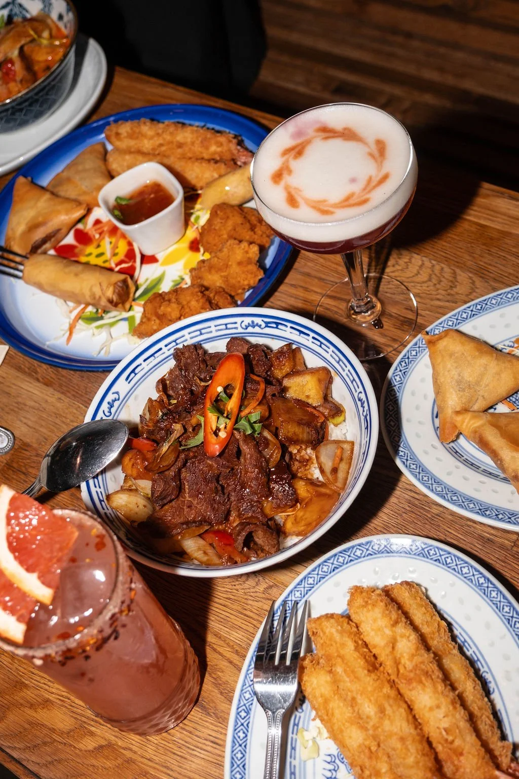 Variety of dishes including fried spring rolls, fried chicken, beef stir-fry, fried fish fillets, and a cocktail with whipped cream and caramel sauce on top, all served on decorative plates on a wooden table.