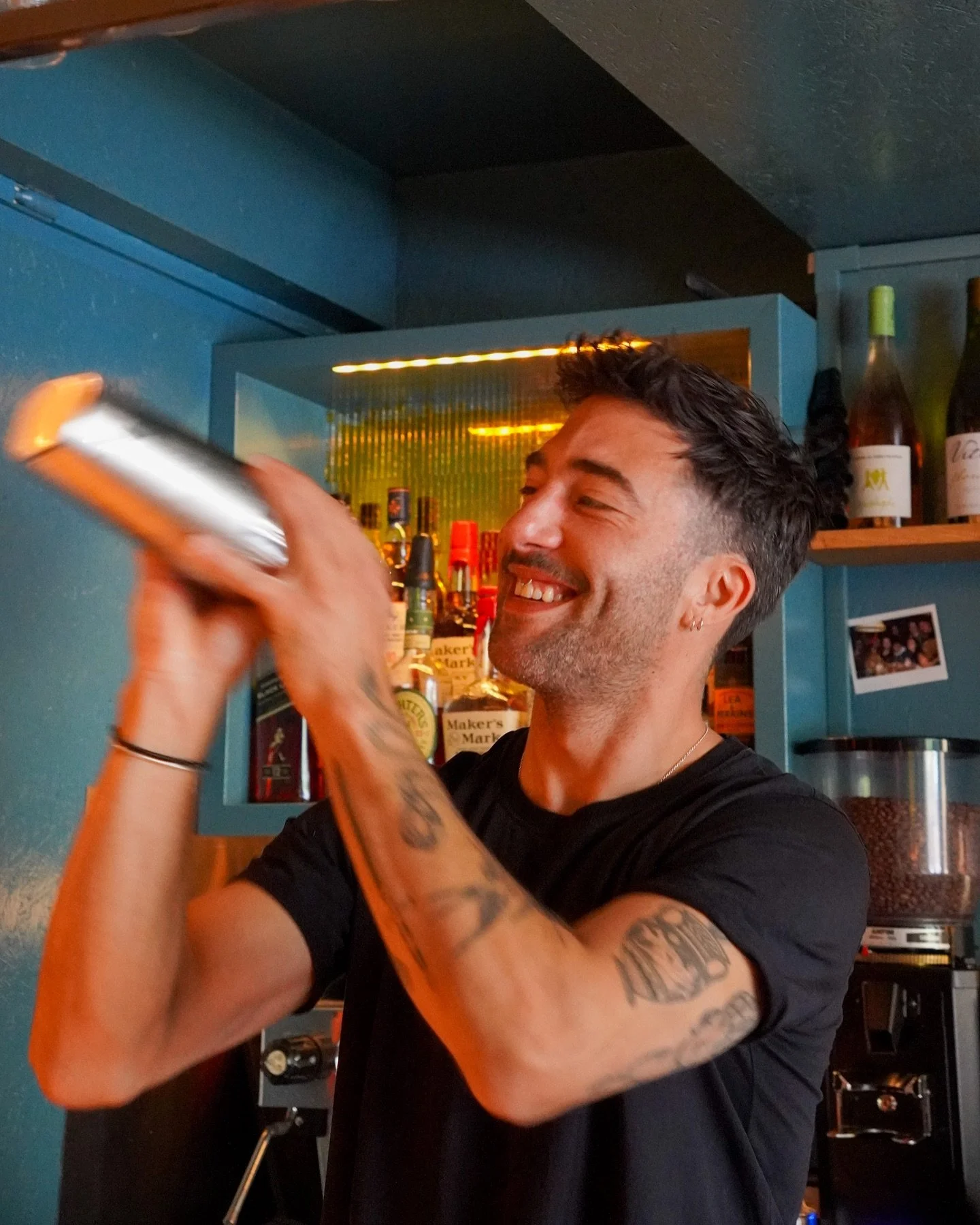 A smiling man with tattoos on his arms is shaking a cocktail shaker behind a bar counter.