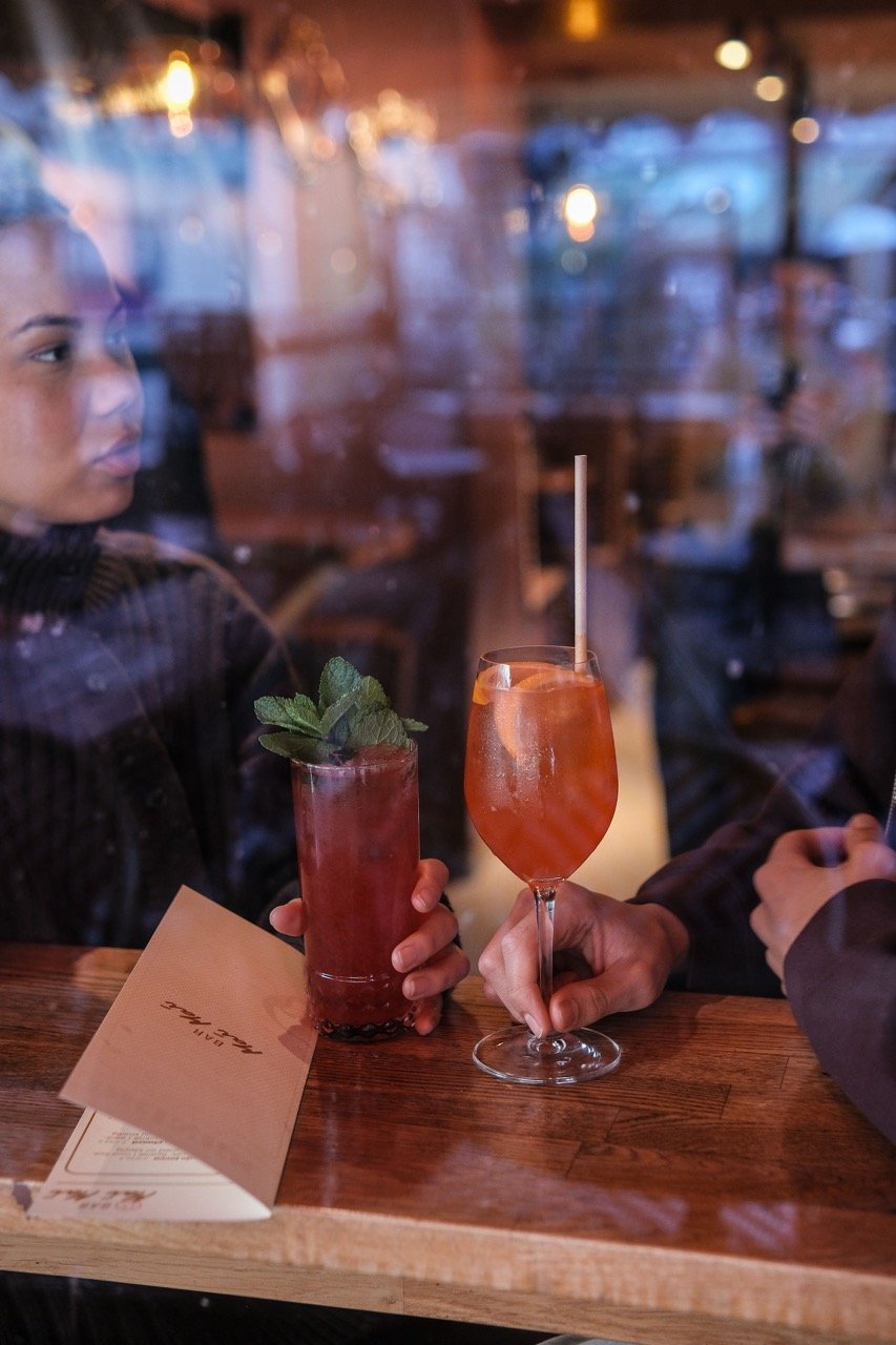 Three cocktails on a wooden table, from left to right: a dark red drink with a foamy top, a tall pink drink with orange slices and salt rim, and a dark coffee-colored drink with cream and coffee beans on top.