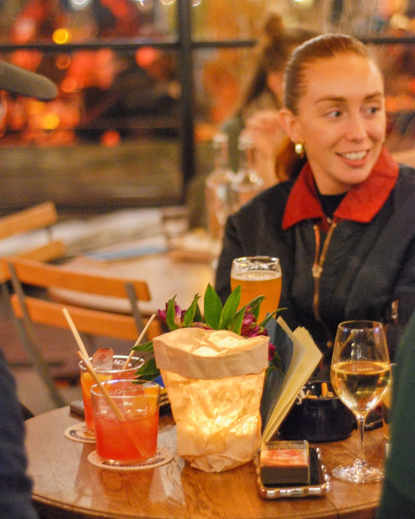 A woman with red hair in a ponytail, wearing a black jacket with a red collar, sitting at a restaurant table with drinks, flowers, and a candle.