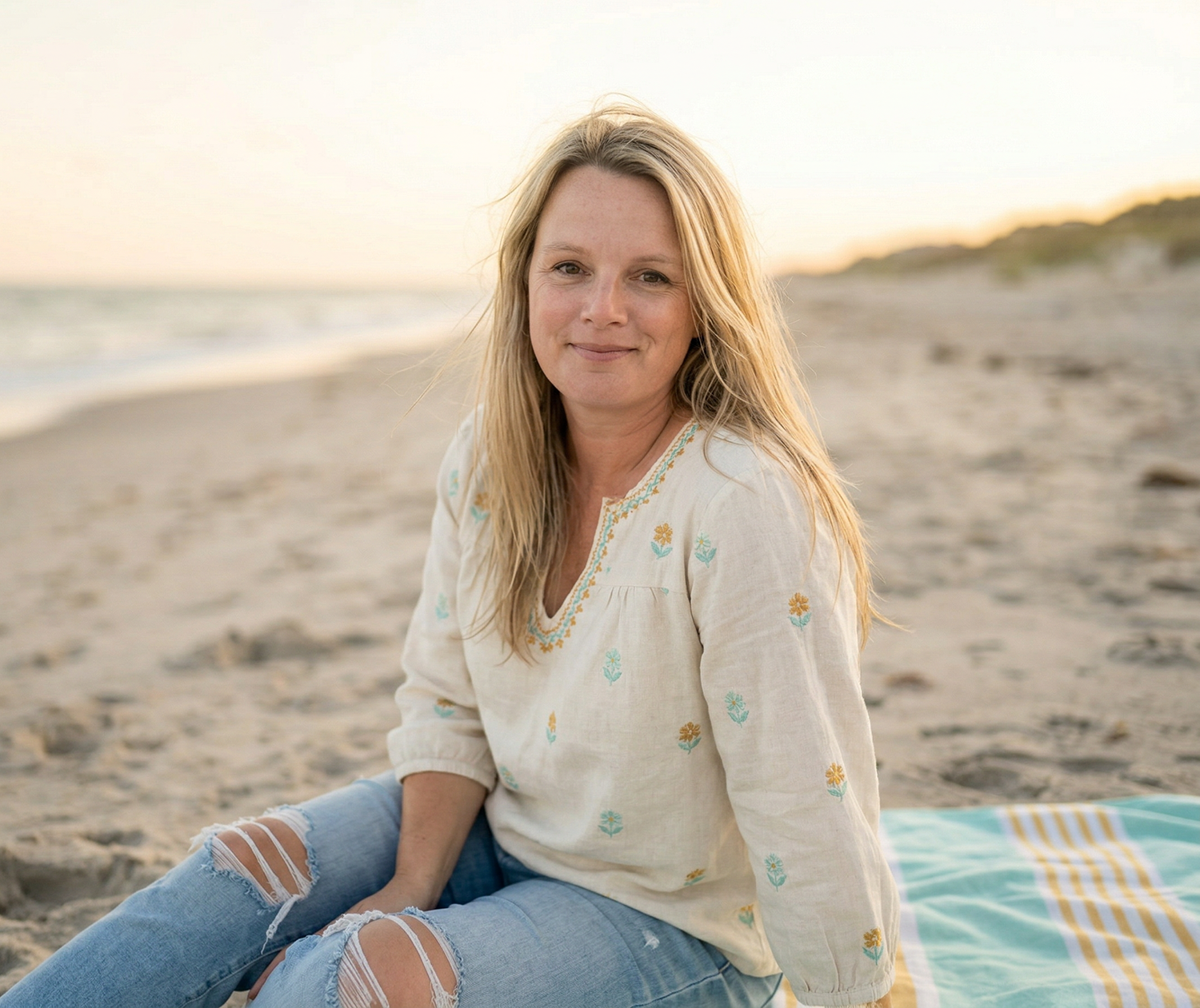 A woman with blonde hair wearing a cream-colored embroidered blouse and ripped jeans sitting on a beach with sand and water in the background during sunset.