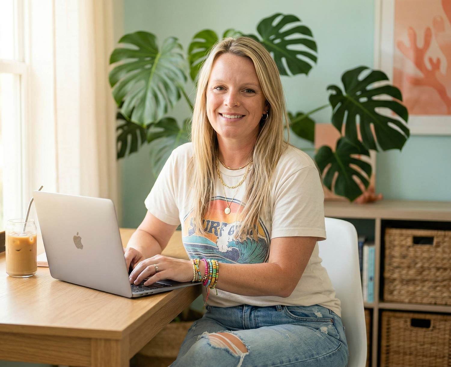 A woman with blonde hair, wearing a graphic T-shirt and ripped jeans, sits at a wooden table with a laptop and iced coffee, smiling at the camera in a room decorated with large green plants and artwork.