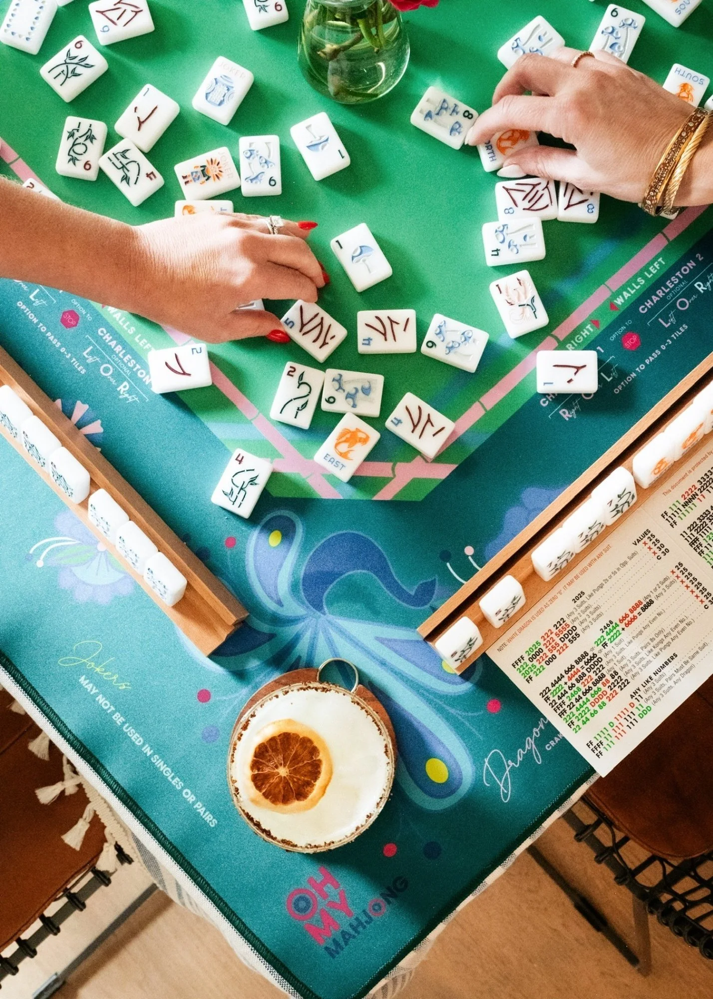 People playing Mahjong, with tiles scattered on a colorful game table that has a butterfly design, and a drink with dried citrus slice nearby.