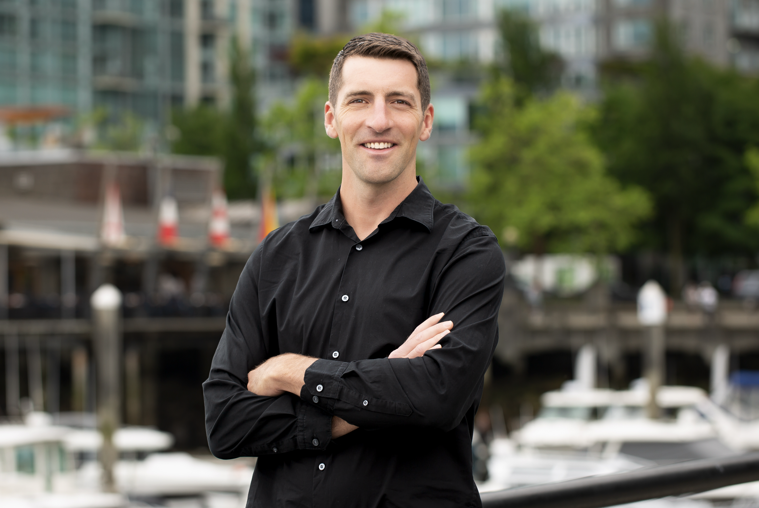A man in a black shirt smiling with arms crossed standing outdoors with blurred buildings and greenery in the background.