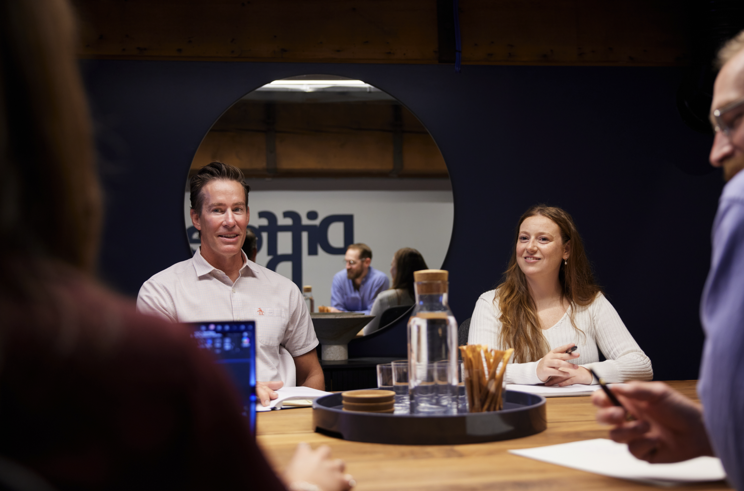People engaged in a conversation at a table in a meeting room, with a circular mirror on the wall reflecting some of the participants and a sign reading 'DIC' in the background.
