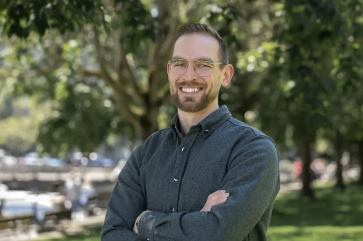 A man with glasses and a beard smiling outdoors in front of trees.