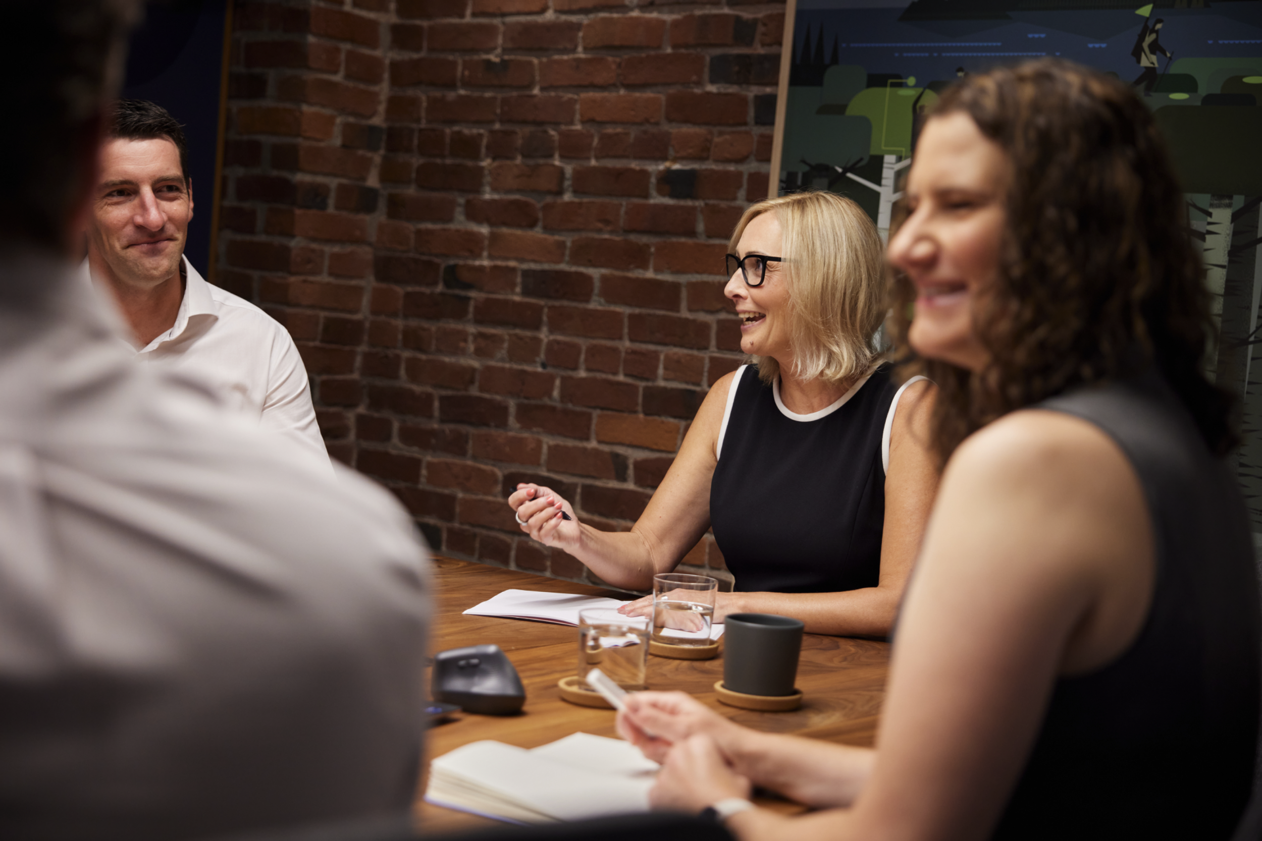 A group of people at a meeting or discussion, sitting around a wooden table in front of a brick wall, engaged and smiling, with notebooks, drinks, and microphones on the table.