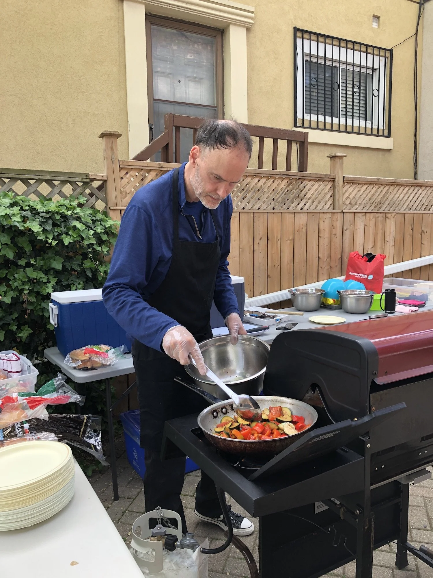 A man grilling vegetables on a barbecue grill outdoors, with a table of food and supplies nearby.