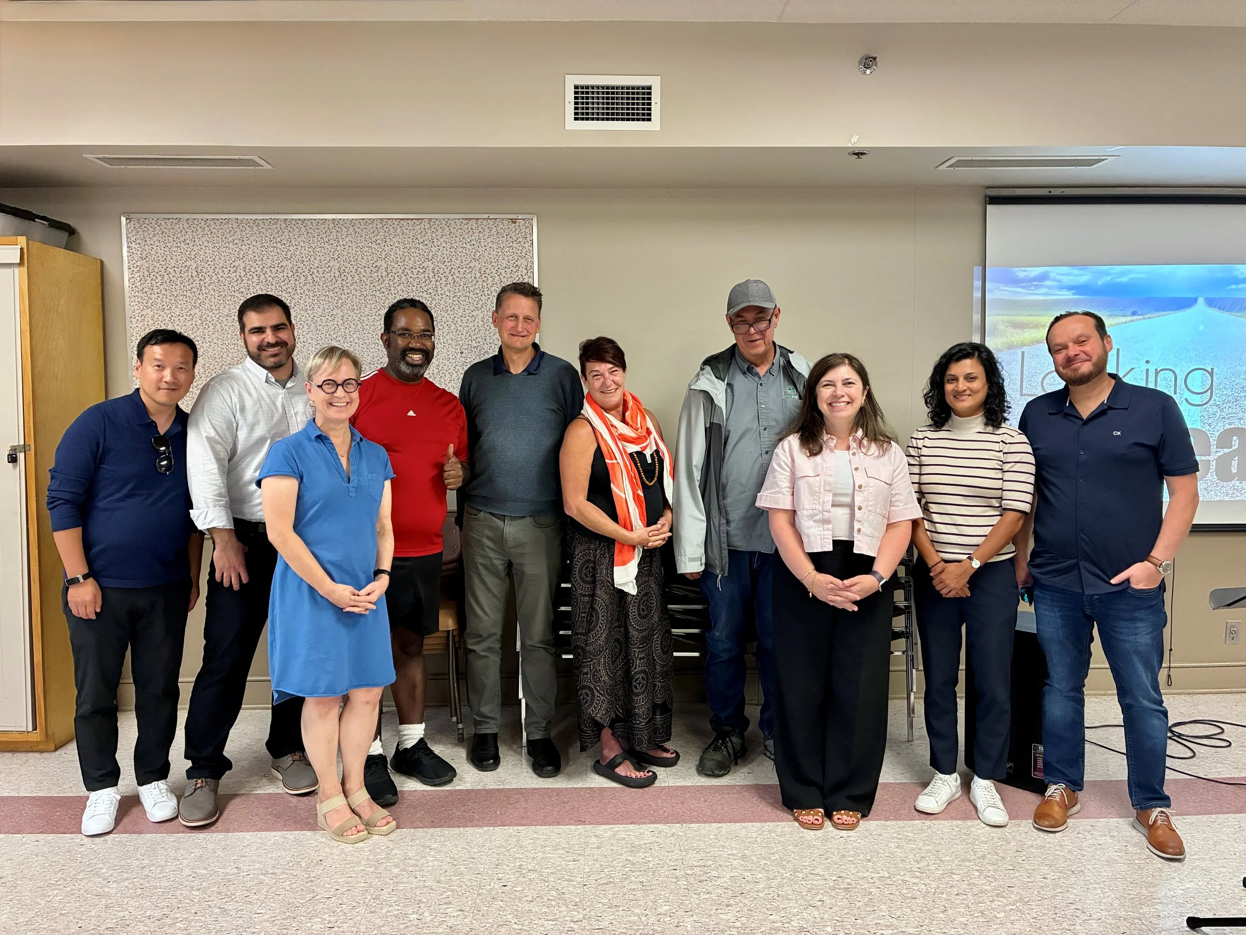 Group of eleven diverse people standing together indoors, smiling for a photo during a presentation or meeting, with a screen in the background displaying the text "Learning"