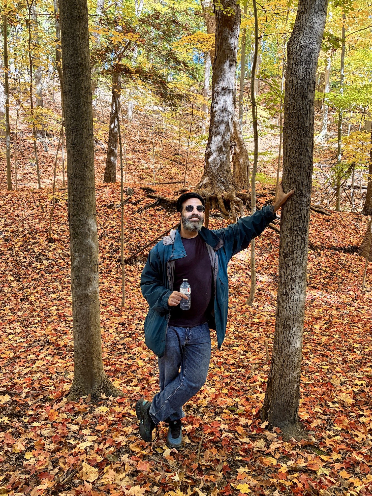 A man with sunglasses, a beard, and a dark jacket smiling and holding a water bottle while standing in a forest with autumn leaves on the ground and tall trees.