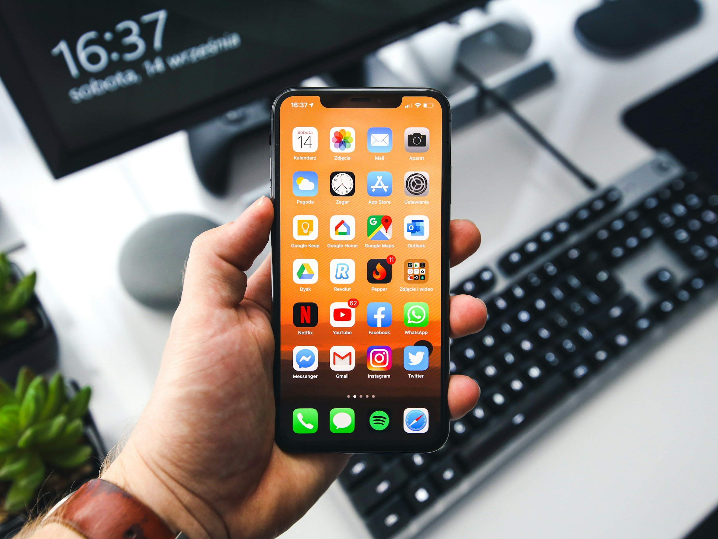 A person holding an iPhone with the home screen showing various app icons, with a computer monitor, keyboard, and a plant in the background.