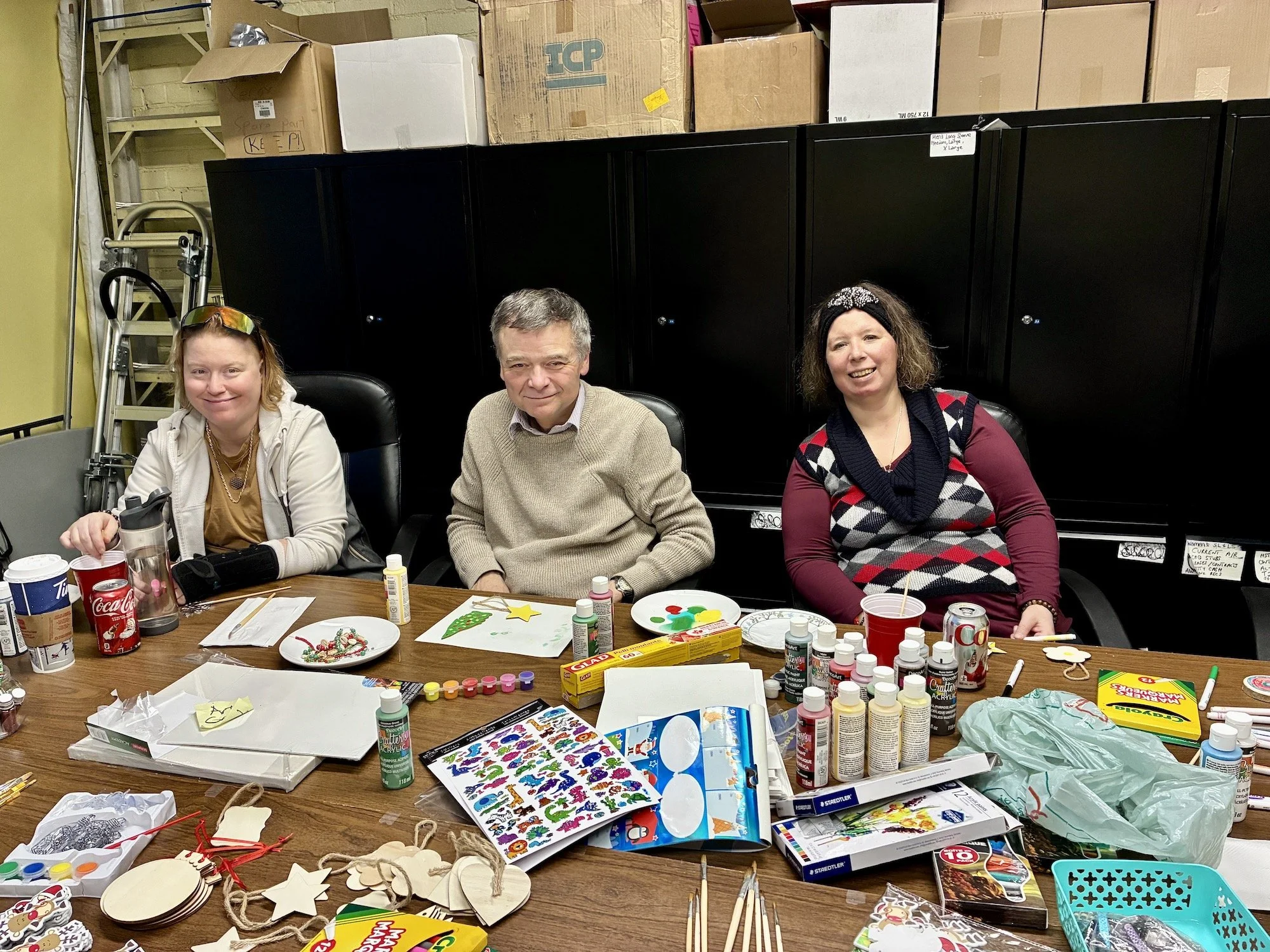 Three women sitting at a table filled with Christmas craft supplies, including paint, stickers, and decorations. The table is in a room with storage cabinets and boxes in the background.