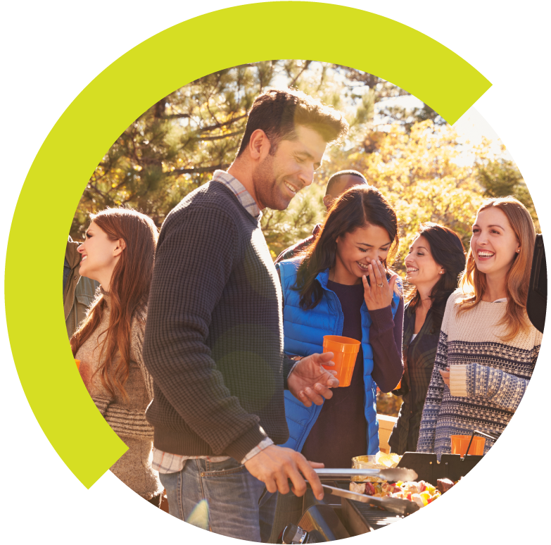 Group of people outdoors enjoying a barbecue, smiling and chatting, with a man grilling food on a barbecue grill.
