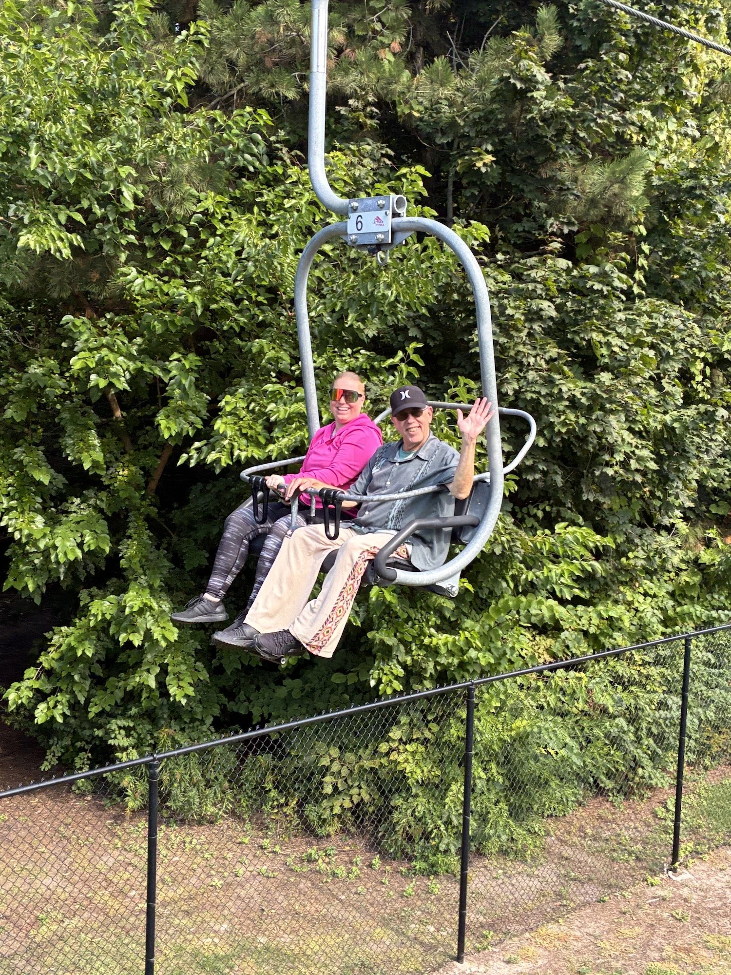 Two people riding a ski lift in a forested area, waving at the camera. The person on the left wears a pink jacket and sunglasses, while the person on the right wears a gray shirt, a black cap, and glasses.
