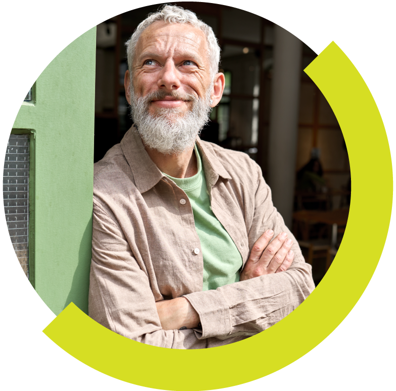 A smiling older man with white hair and a beard, wearing a light brown shirt and green T-shirt, standing with arms crossed in a room with green walls and natural light.