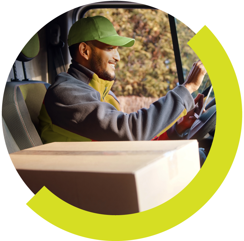A smiling man wearing a green cap and gray jacket sitting in a delivery truck, holding a smartphone with a cardboard box in front of him, outdoors with trees in the background.