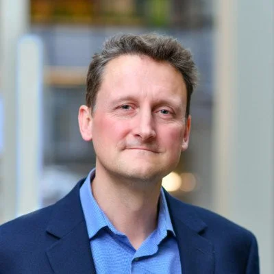 A man with short brown hair wearing a blue dress shirt and dark blazer, standing in front of a blurred background.