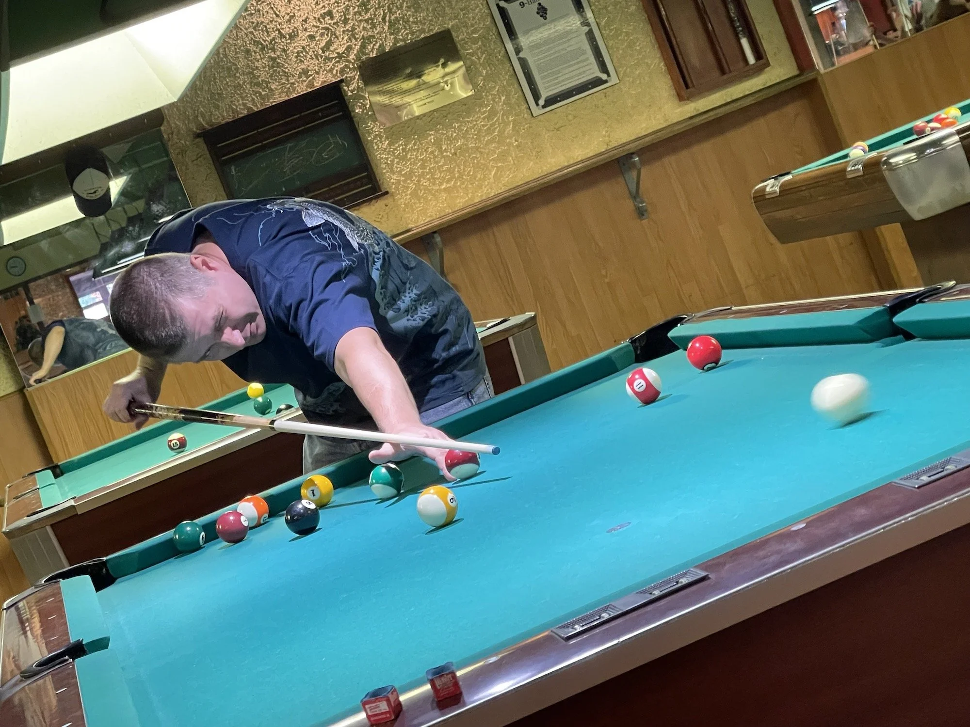 A man playing pool at a billiards table, lining up a shot with a cue stick surrounded by pool balls on the table in a room with wood-paneled walls and mirrors.