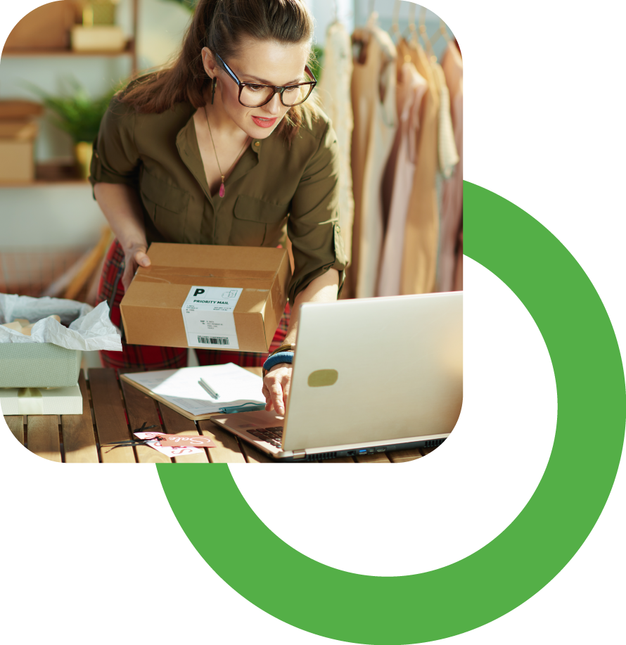 A woman with glasses and brown hair, wearing a green shirt, pulls a small cardboard box with a shipping label from a desk with a laptop, papers, and packaging in a workshop or retail setting with hanging clothes in the background.
