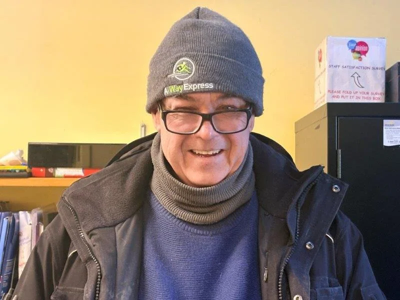 A smiling man wearing glasses, a gray beanie with a sports logo, a gray scarf, and a dark jacket, standing indoors near a yellow wall and black shelves with boxes and books.