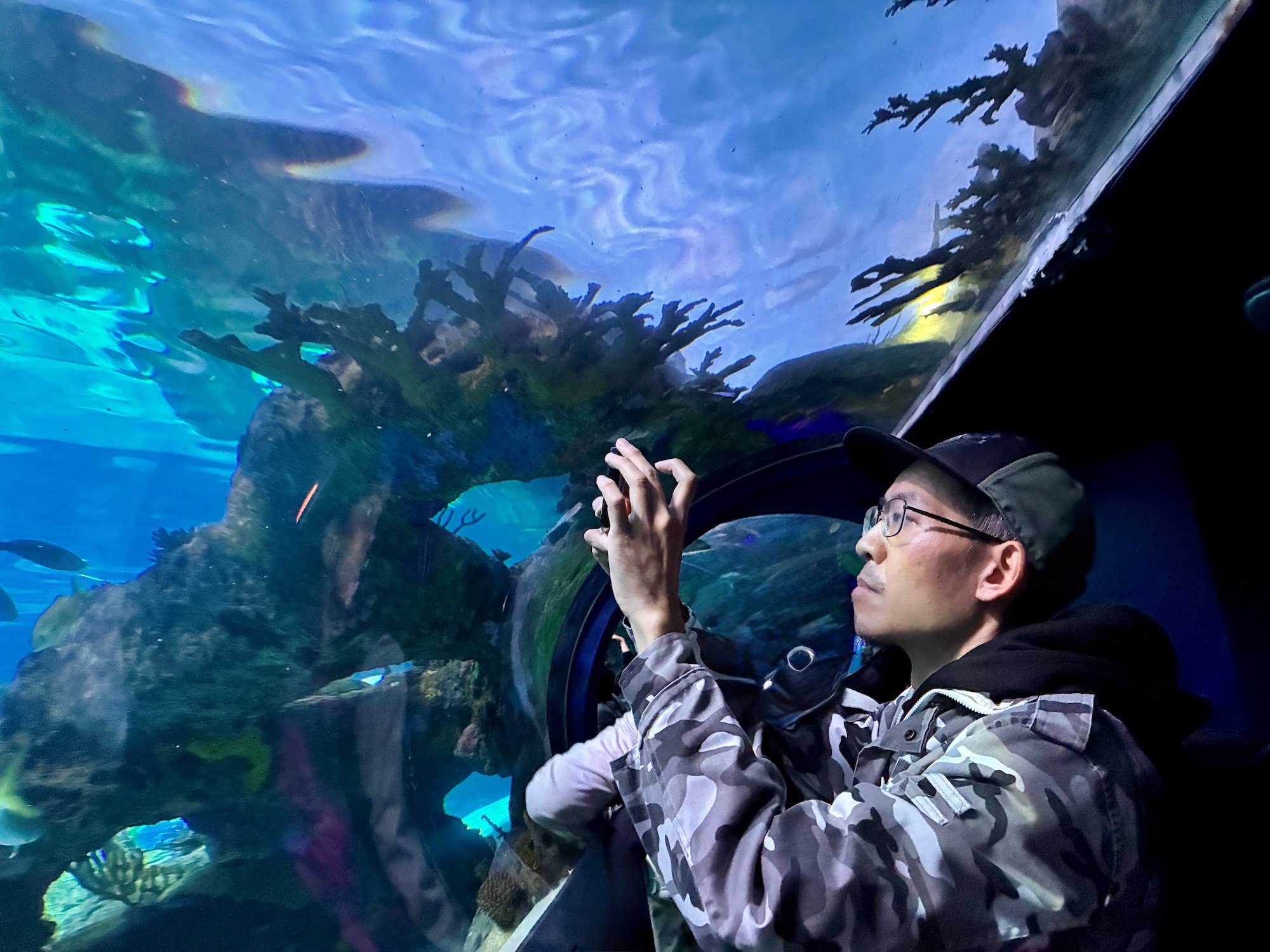 Man in camouflage jacket and glasses taking a photo through an aquarium window with colorful fish and coral in the water.