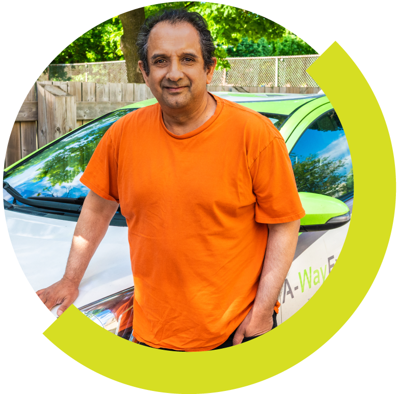 Man standing outdoors next to a white and green electric vehicle, wearing an orange t-shirt, with lush green trees and a wooden fence in the background.