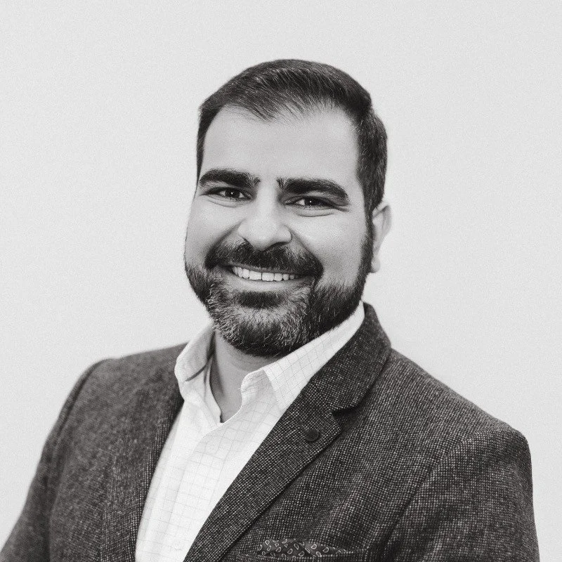 Portrait of a man with dark hair and beard, smiling, wearing a suit jacket and collared shirt against a plain background.